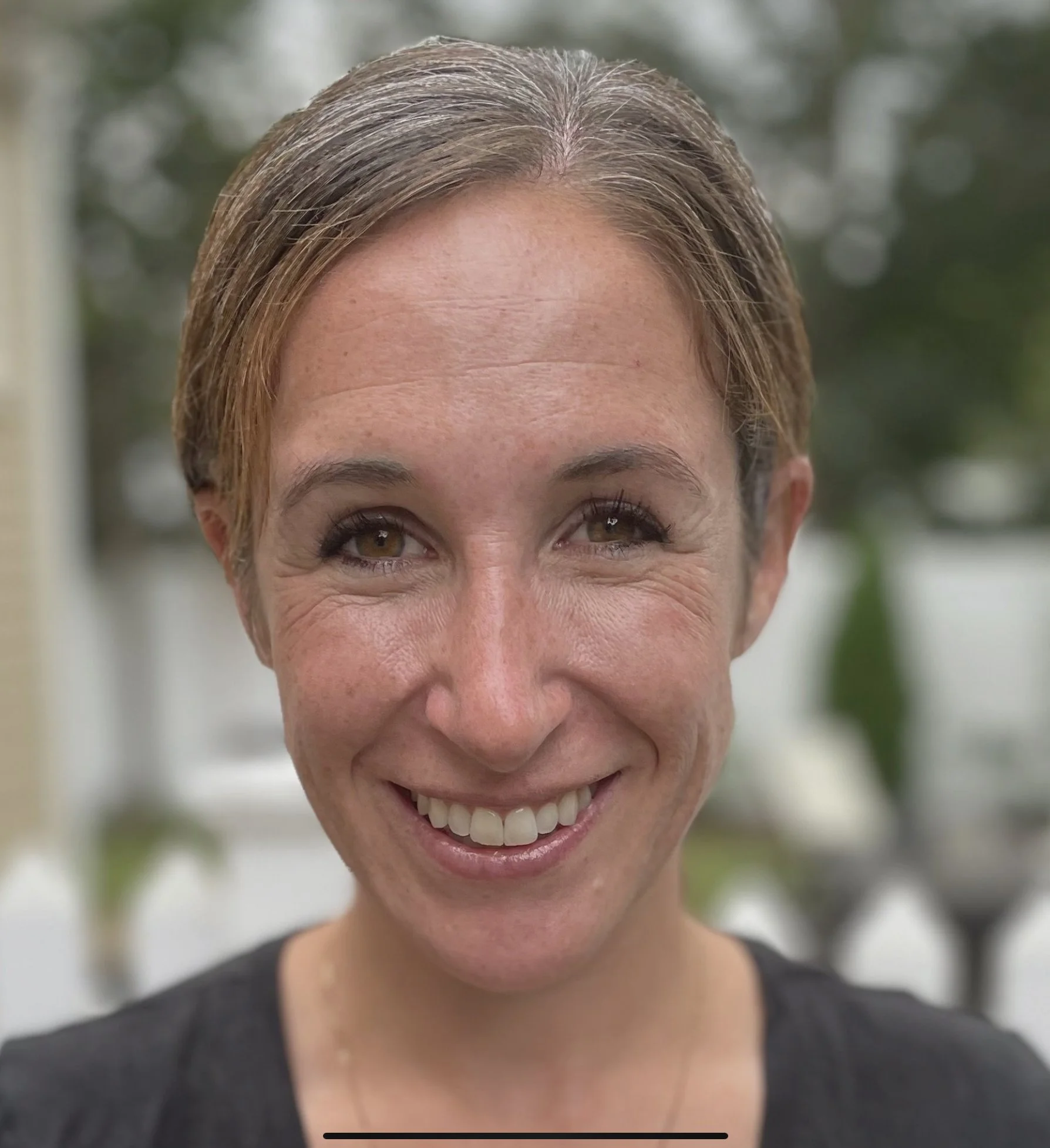 A smiling woman with short hair in an outdoor setting with greenery in the background.