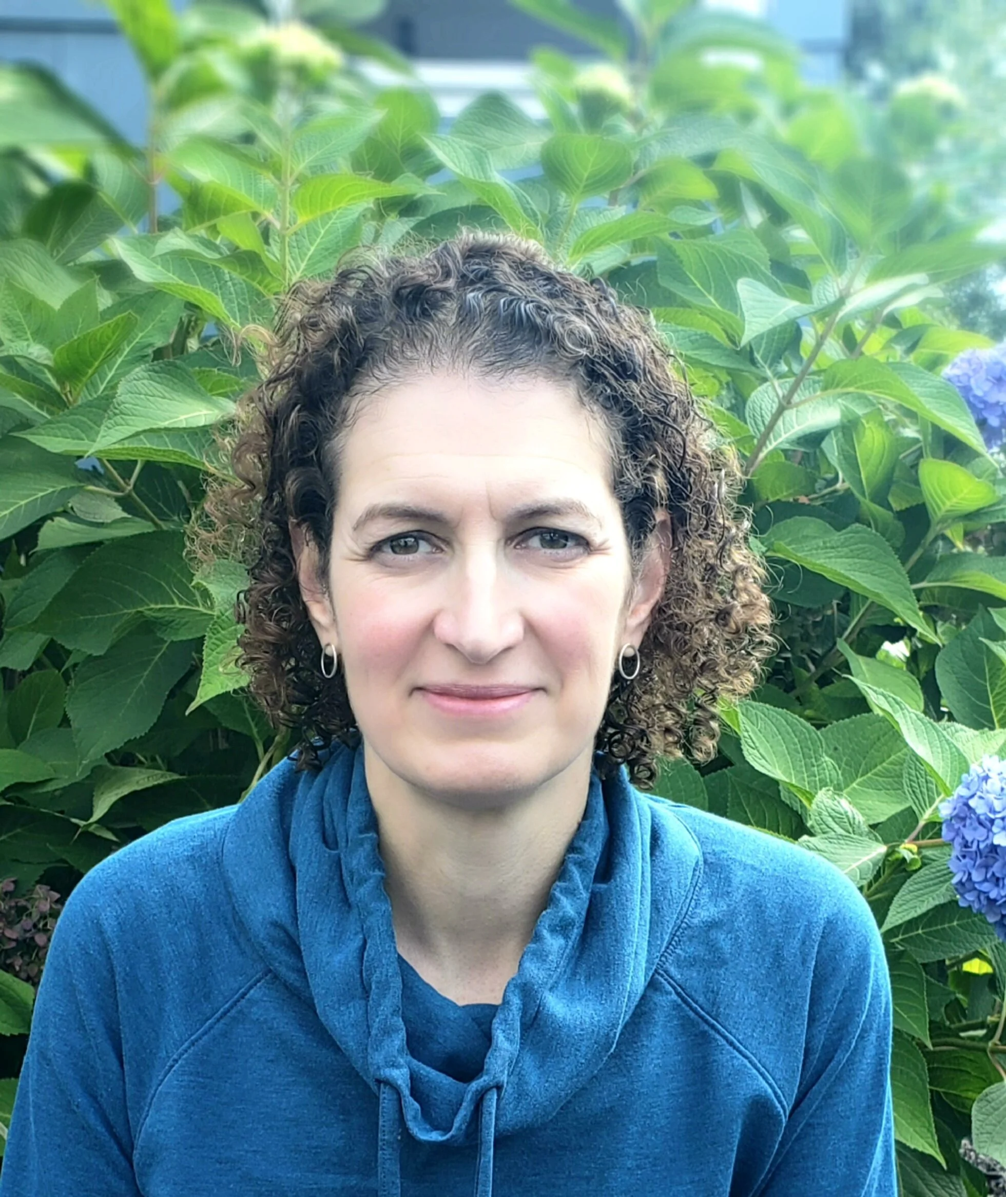 Person with curly hair wearing a blue hoodie standing in front of green foliage and purple flowers.