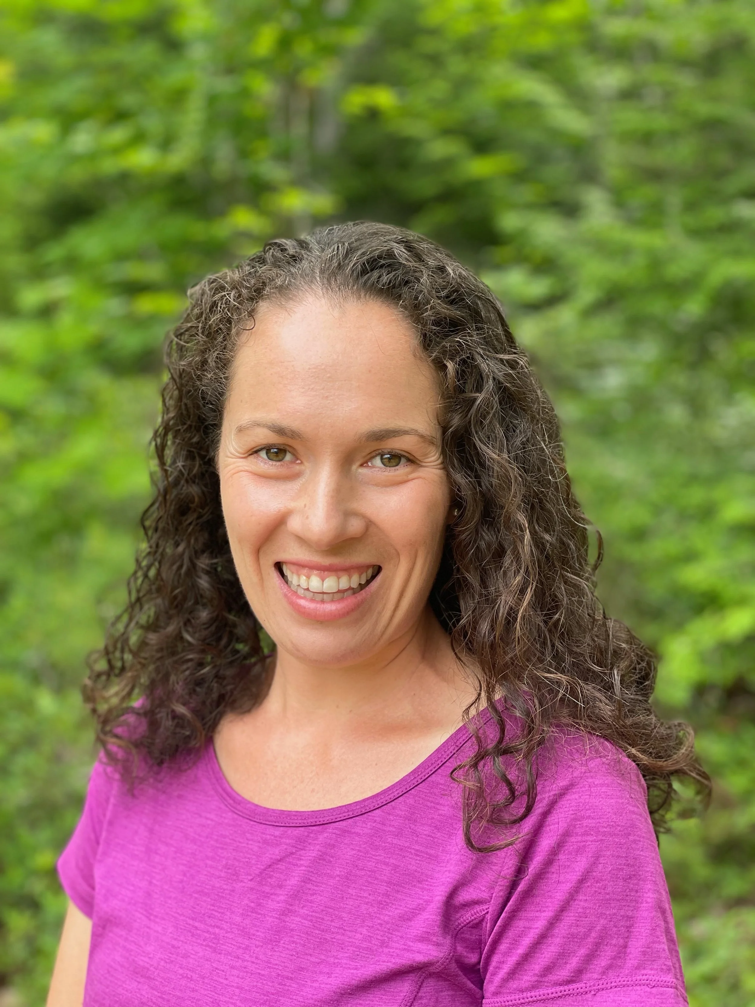 Smiling woman with curly hair wearing a purple shirt in a green outdoor setting.