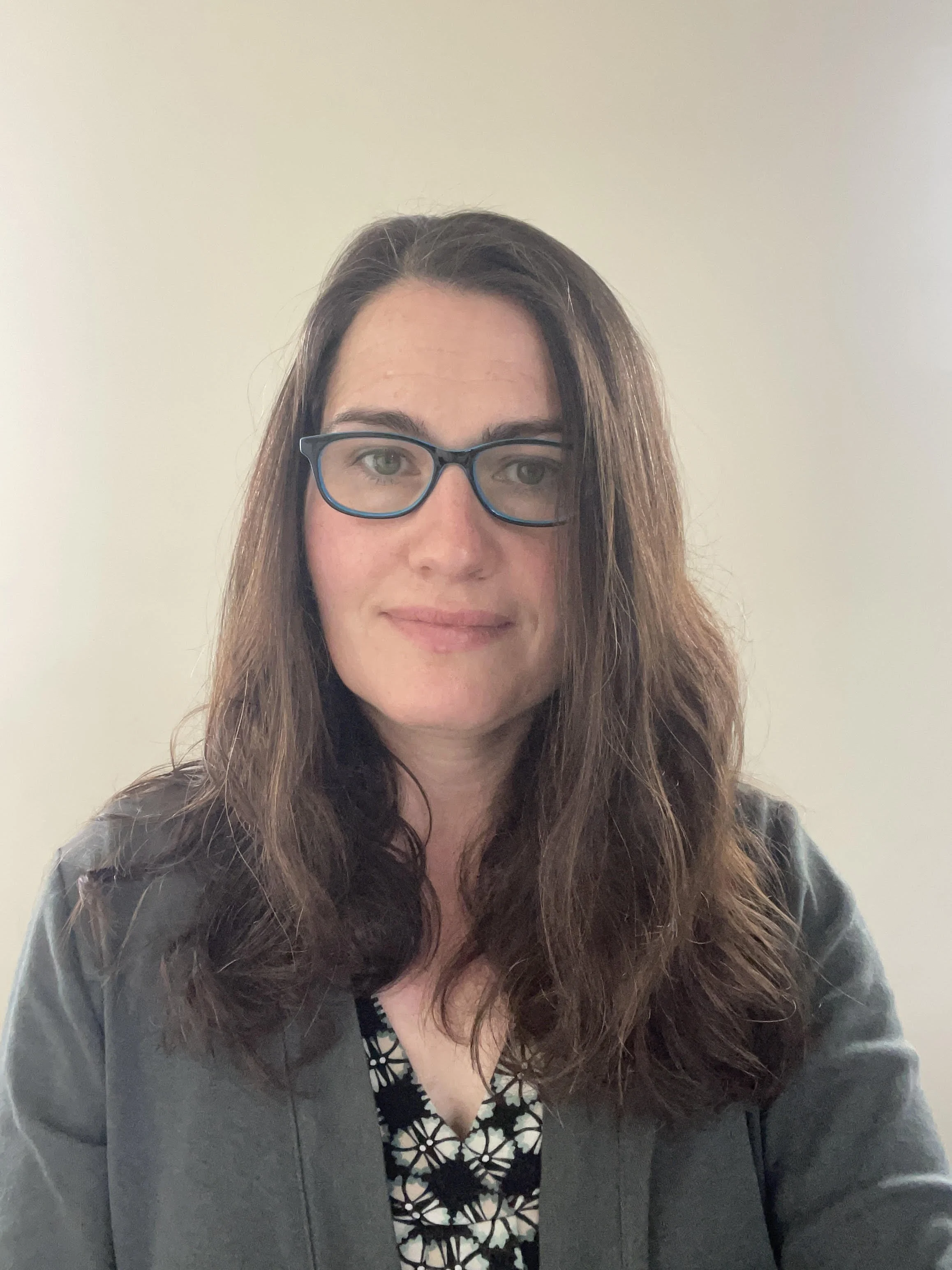 Woman with long brown hair and glasses wearing a dark gray jacket and a patterned top, standing against a plain background.