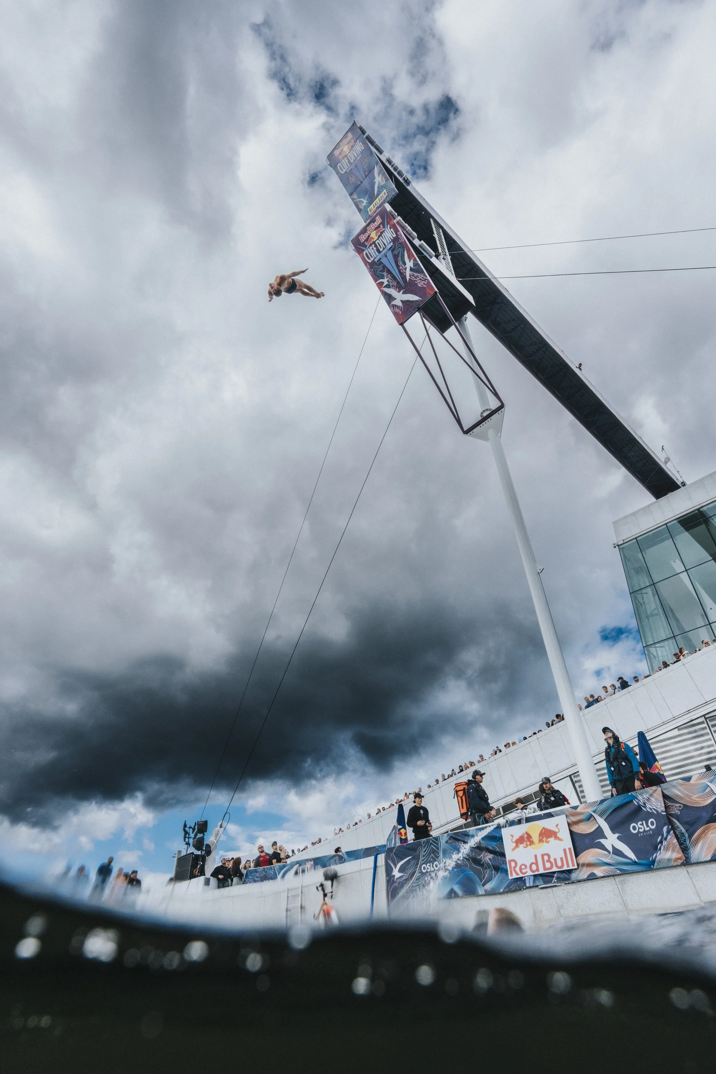 A person diving from a high platform at a Red Bull cliff diving event in Oslo, with spectators watching from a building and a cloudy sky overhead.