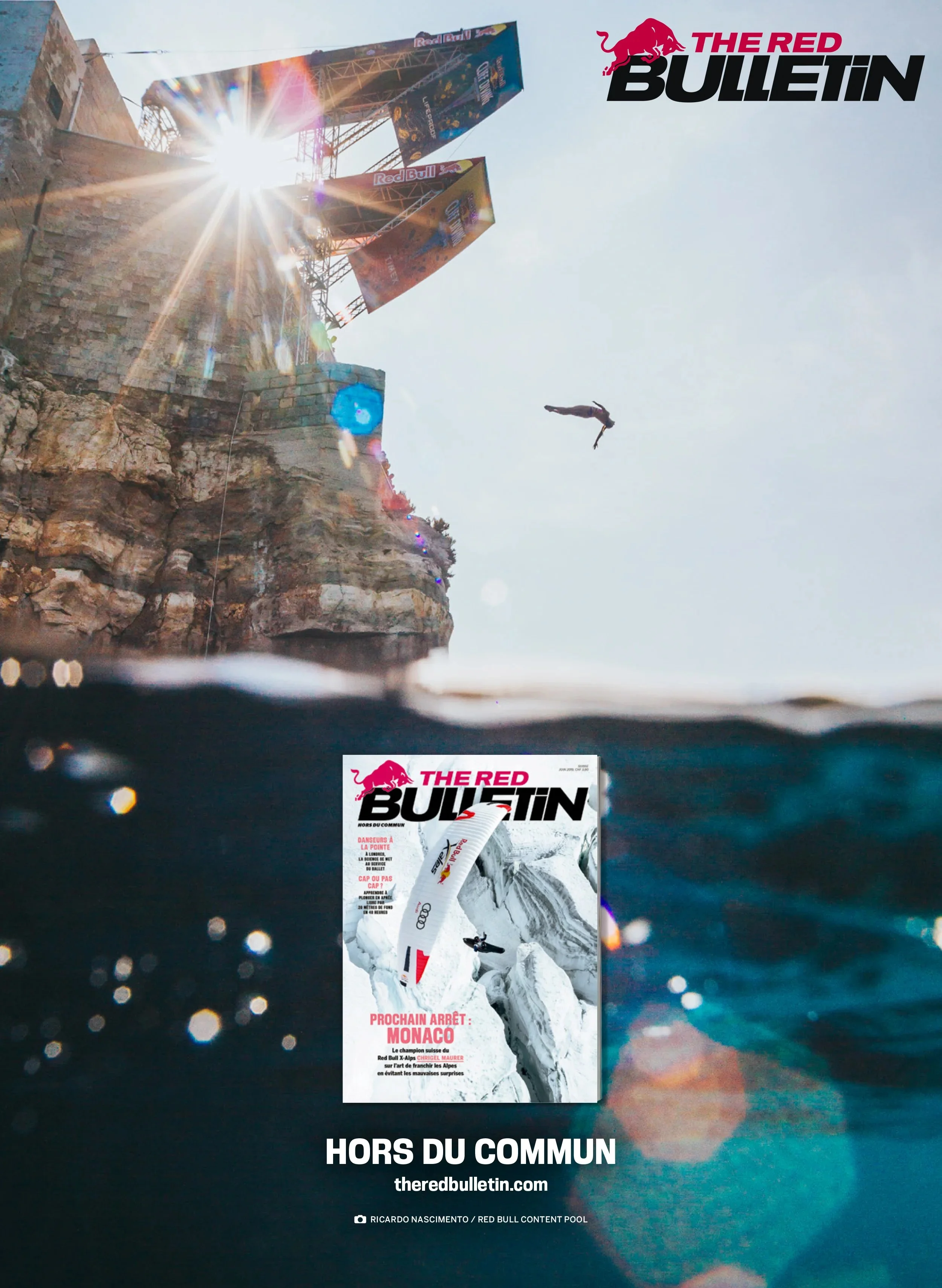 A cliffside with a diving platform and colorful banners, sun shining through the sky, a person diving into the water, and a magazine cover titled 'The Red Bulletin' submerged partially underwater.