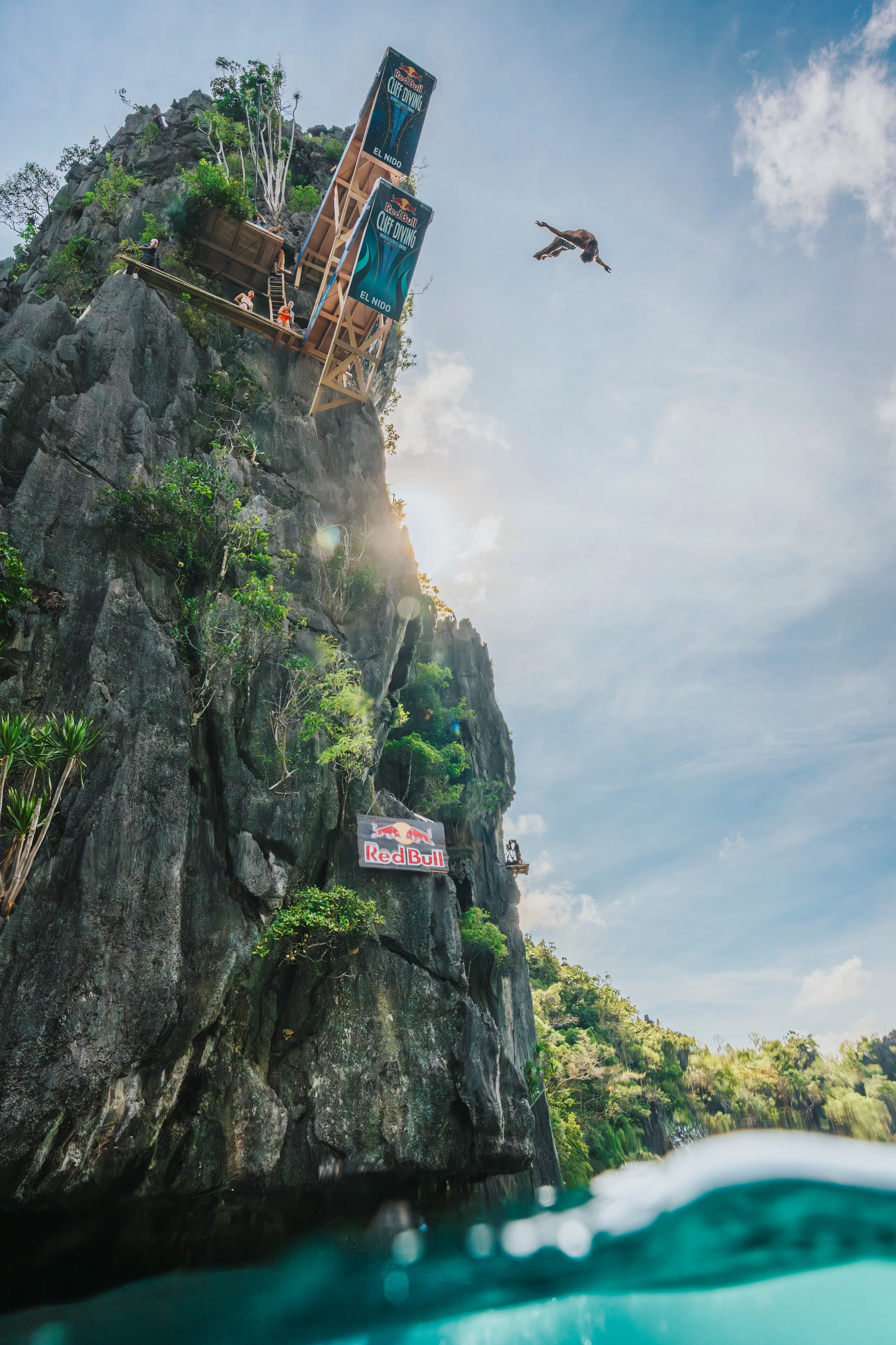 Person midair diving off a cliff at Red Bull Cliff Diving competition in El Nido, with onlookers watching from a platform and a sign on the cliff, against a bright sky.
