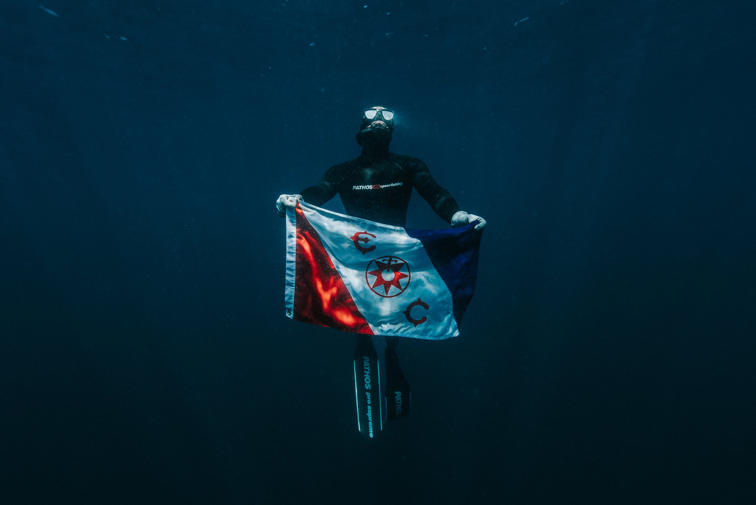 A scuba diver underwater holding a flag with a red, white, and blue design and symbols in the center, wearing a black suit, goggles, and fins.