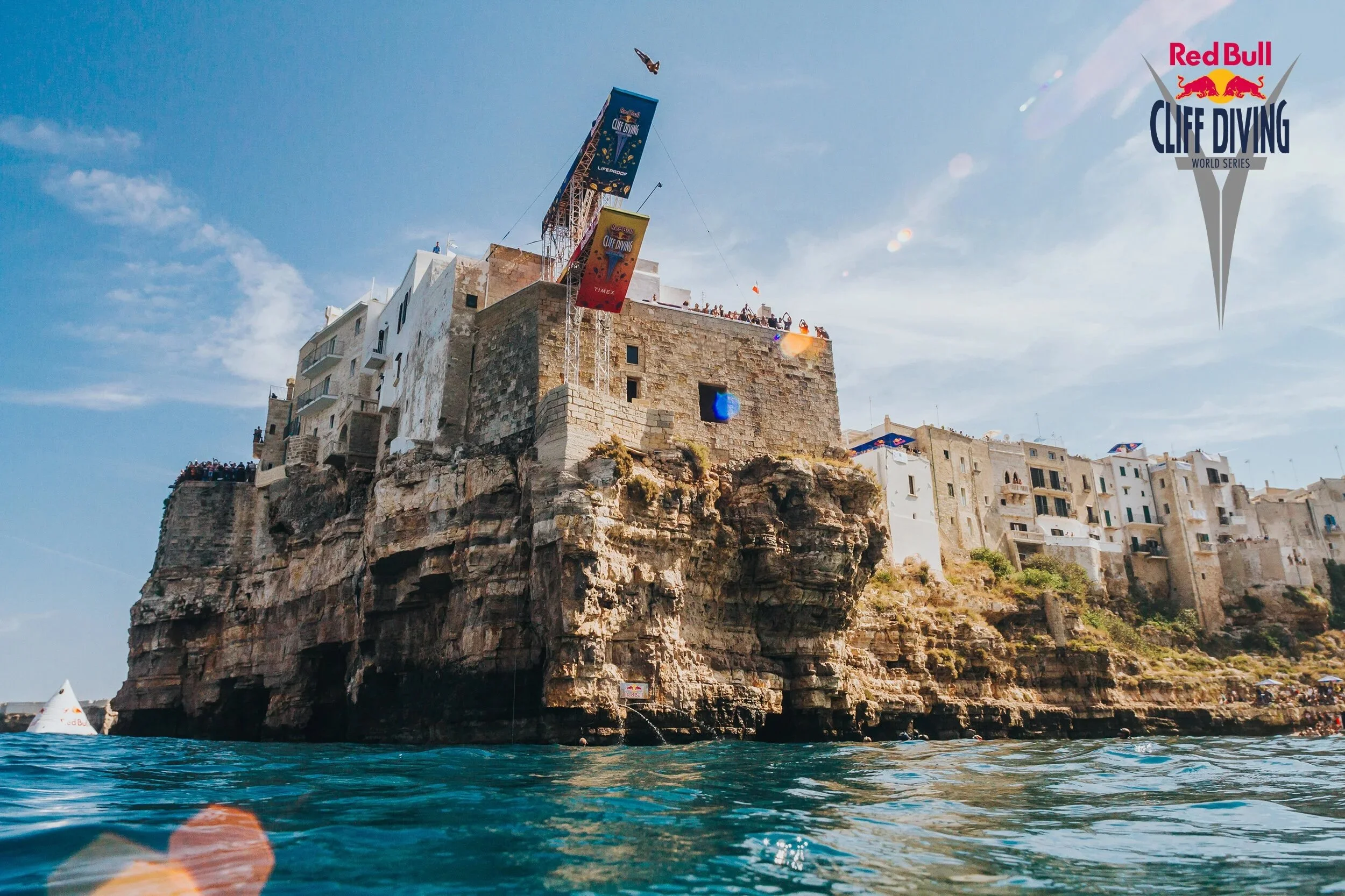 Cliffside buildings with banners for Red Bull Cliff Diving World Series, viewed from the water under a blue sky.