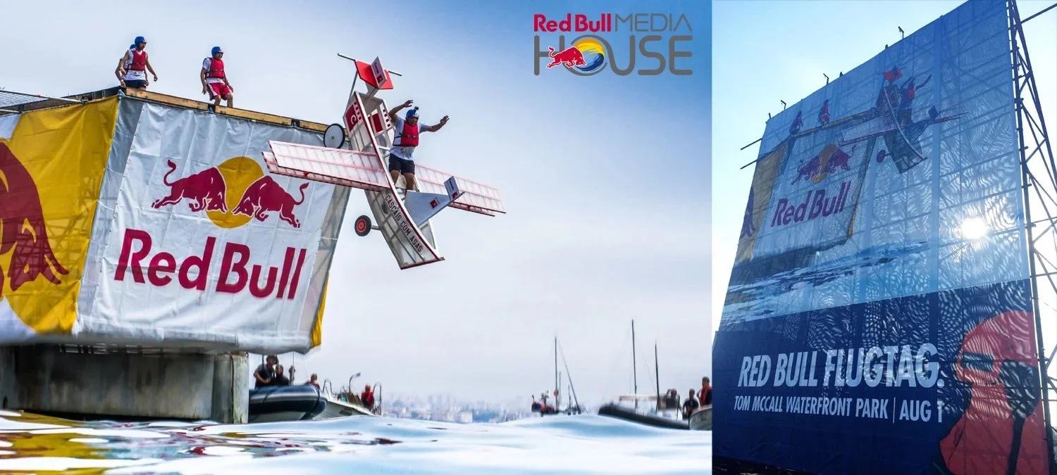 People holding onto a Red Bull themed watercraft in a water-based stunt event. A large banner with the Red Bull logo is visible, and part of a building or platform displaying Red Bull advertising.