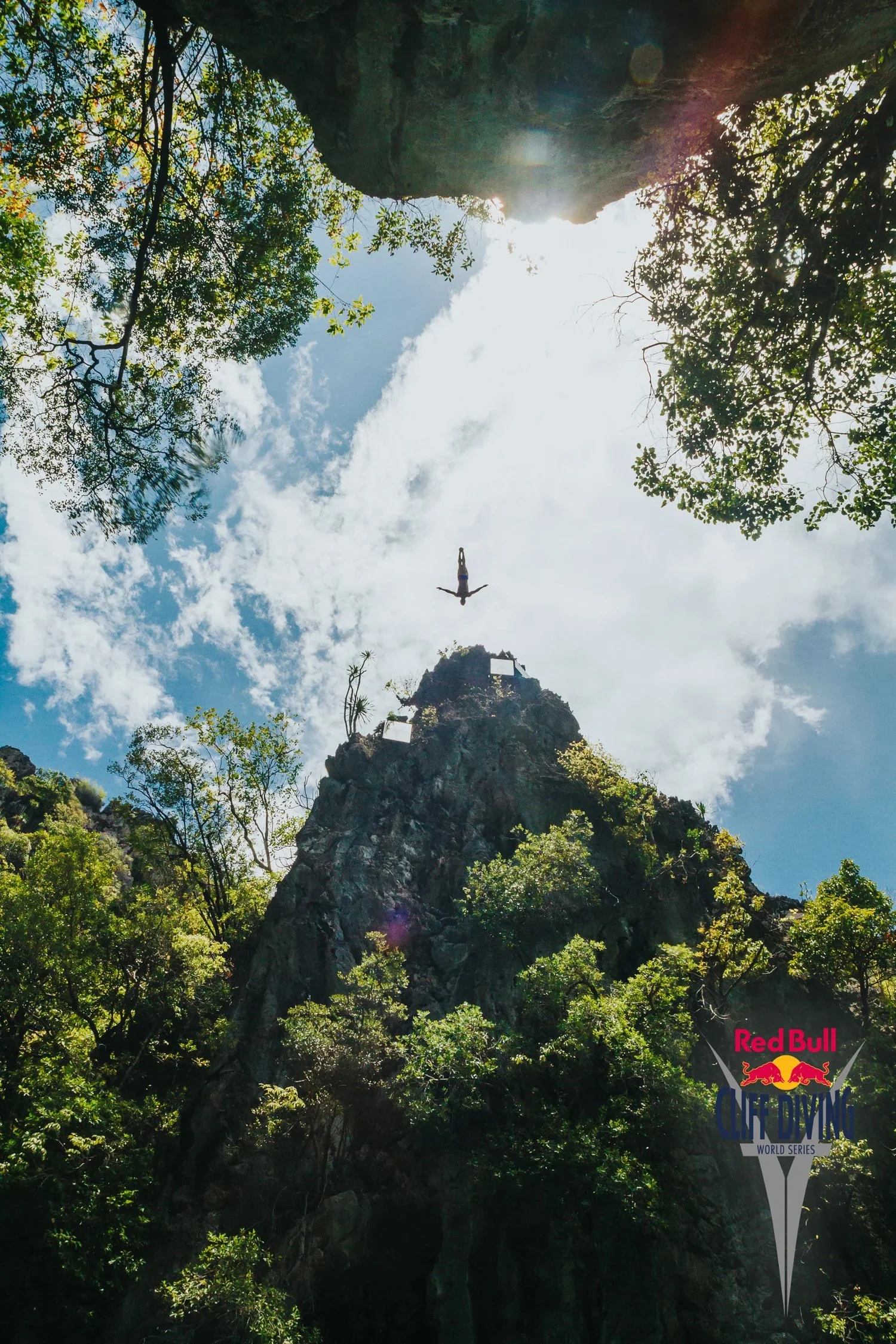 A rock climber is mid-air jumping off a tall, steep rock formation surrounded by trees, with a partly cloudy sky in the background, at the Red Bull Cliff Diving World Series event.