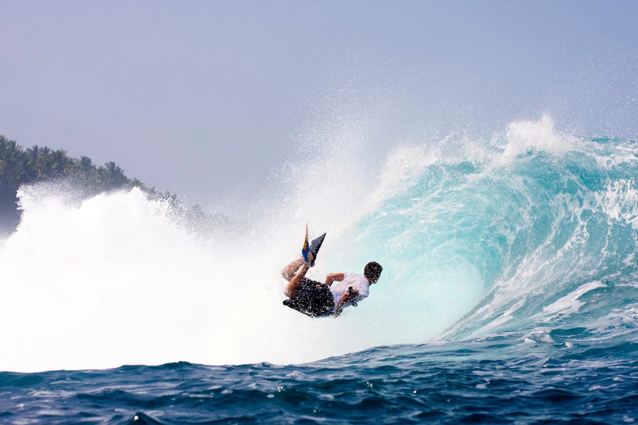 A person surfing on a large ocean wave near a tropical island with palm trees in the background.