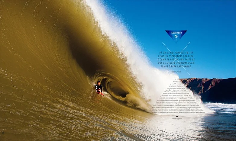 A surfer rides inside a large, yellowish-green wave near a rocky coastline with clear blue sky.