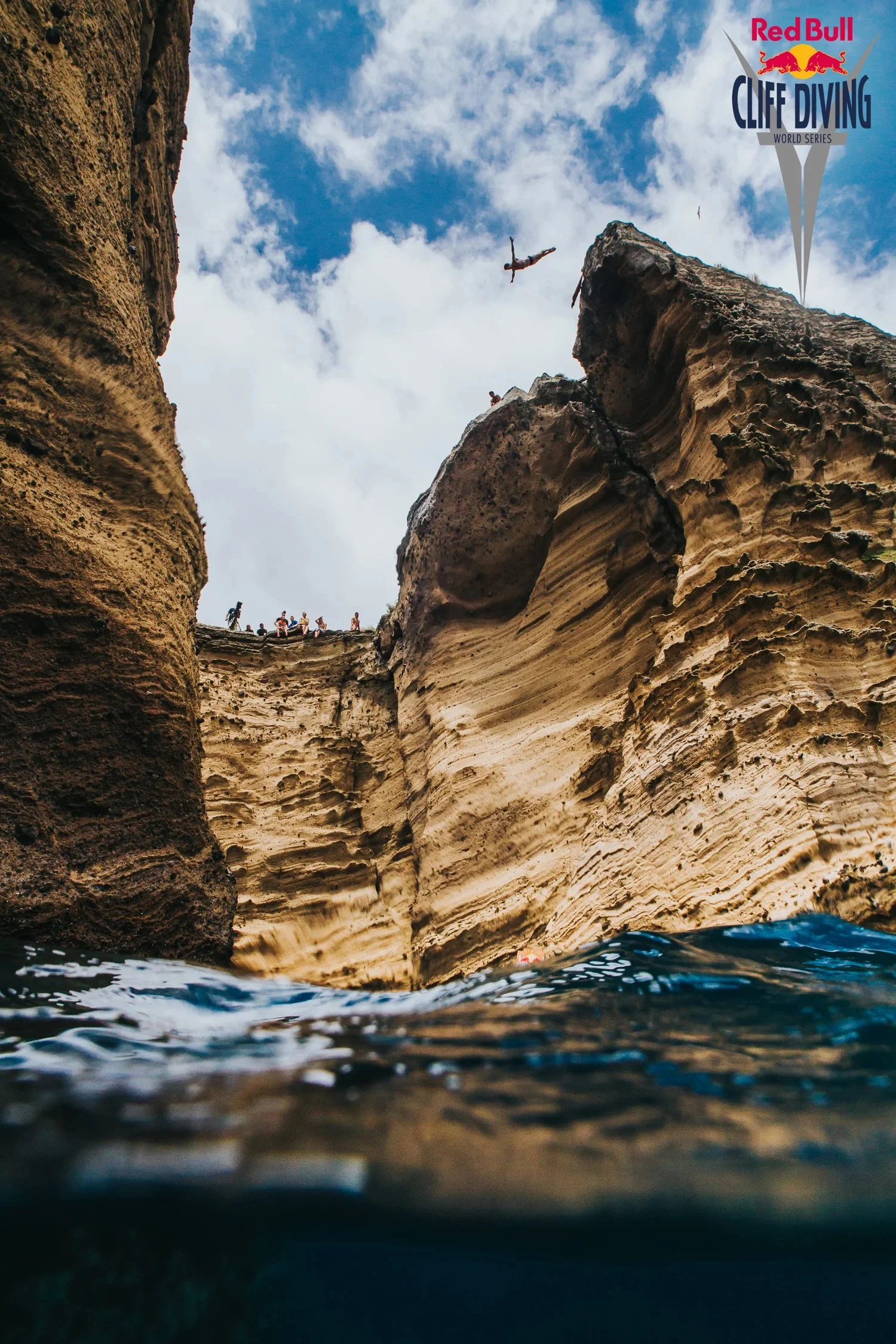 A cliff diver mid-air above a rocky coastal cove with a group of people standing on the edge, blue sky with clouds, and the Red Bull Cliff Diving World Series logo in the top right corner.
