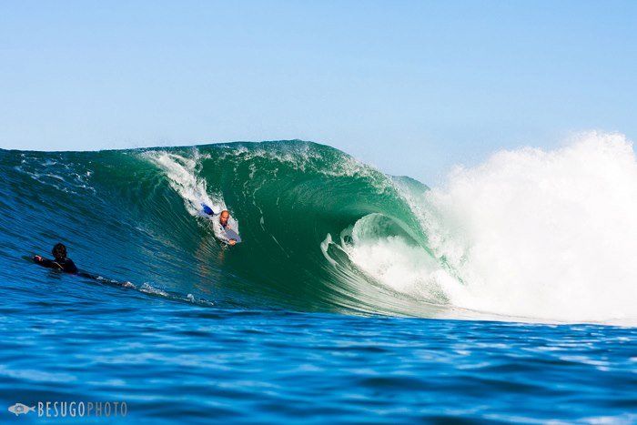 A surfer riding inside the barrel of a large ocean wave while another person watches from the water.