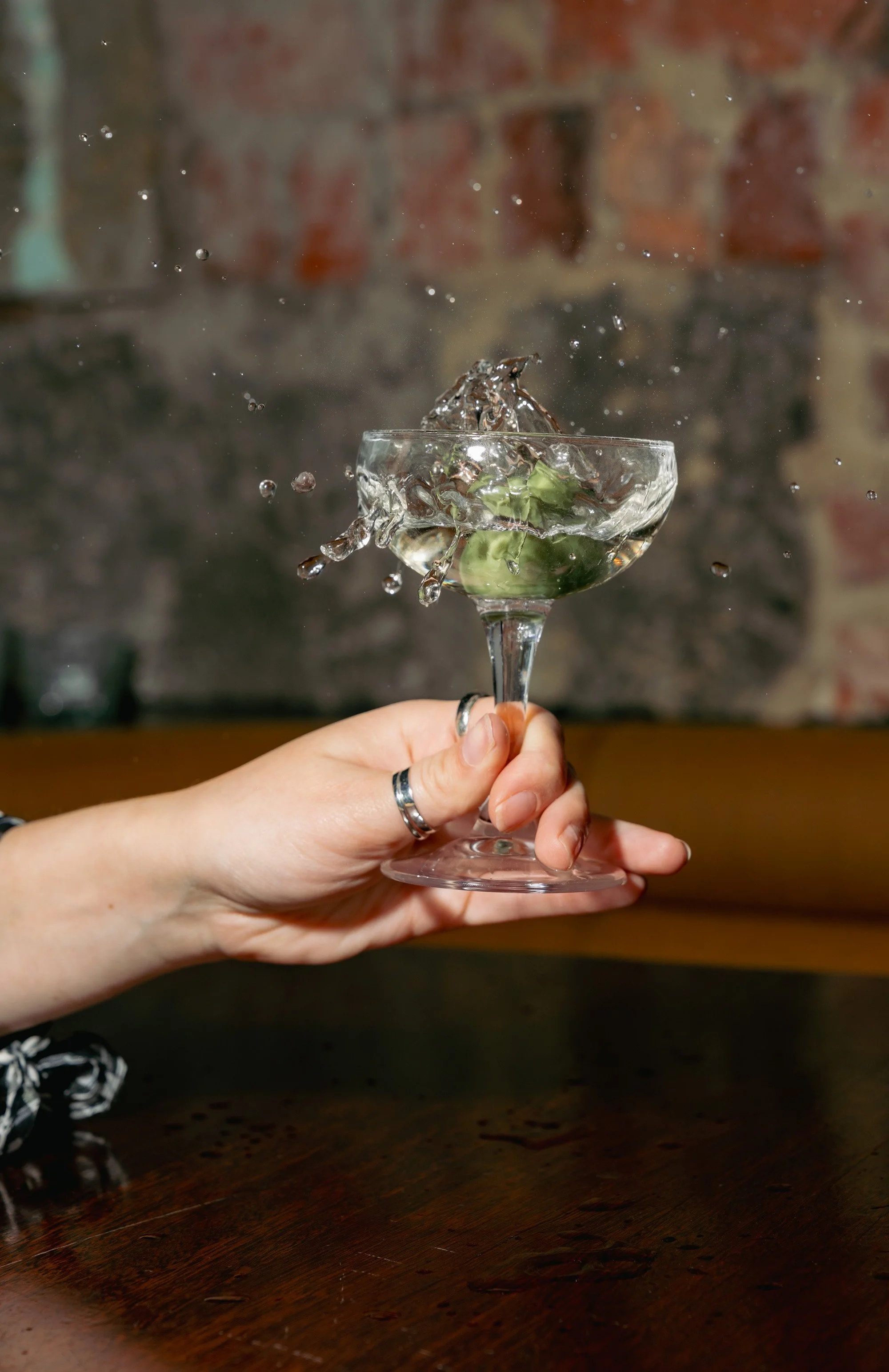 A person holding a clear cocktail glass with an olive inside, causing water to splash out as an ice cube is dropped into it, against a blurred brick wall background.