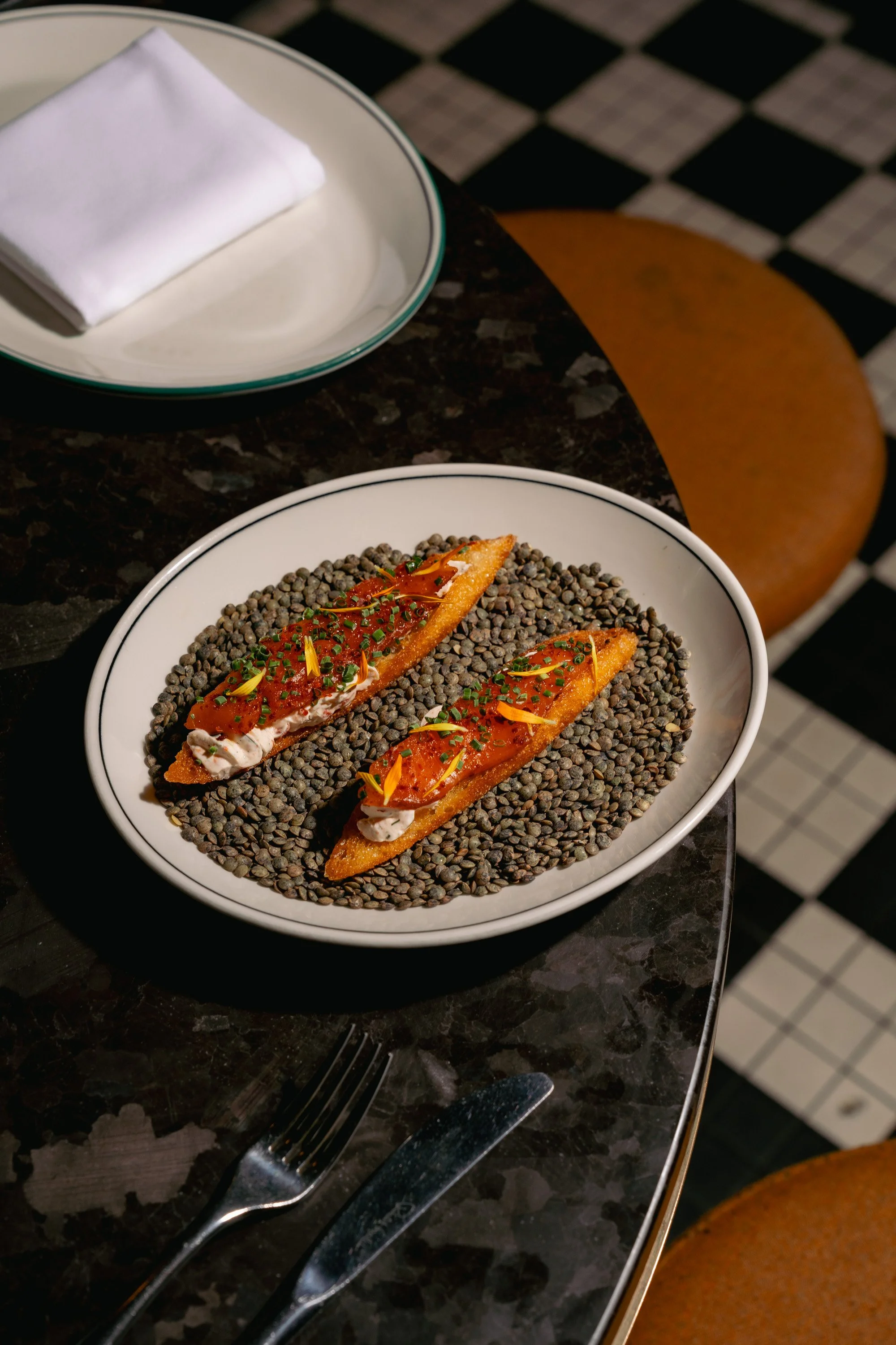 A white bowl with two tostadas topped with shrimp, red sauce, chopped herbs, and edible flower petals, placed on a bed of black lentils. In the background, there is a green plate with a folded white napkin, black fork, and knife on a dark marble tabl
