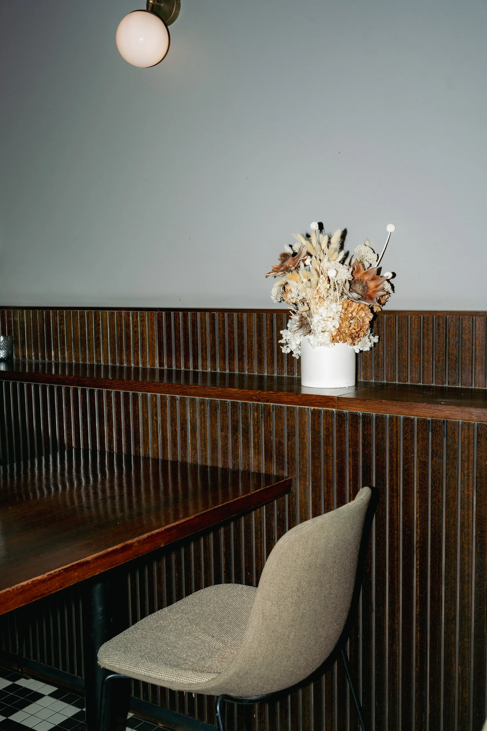 A beige upholstered chair sits next to a wooden table against a wall, with a white vase of dried flowers on a wooden counter, and a wall-mounted light fixture above.