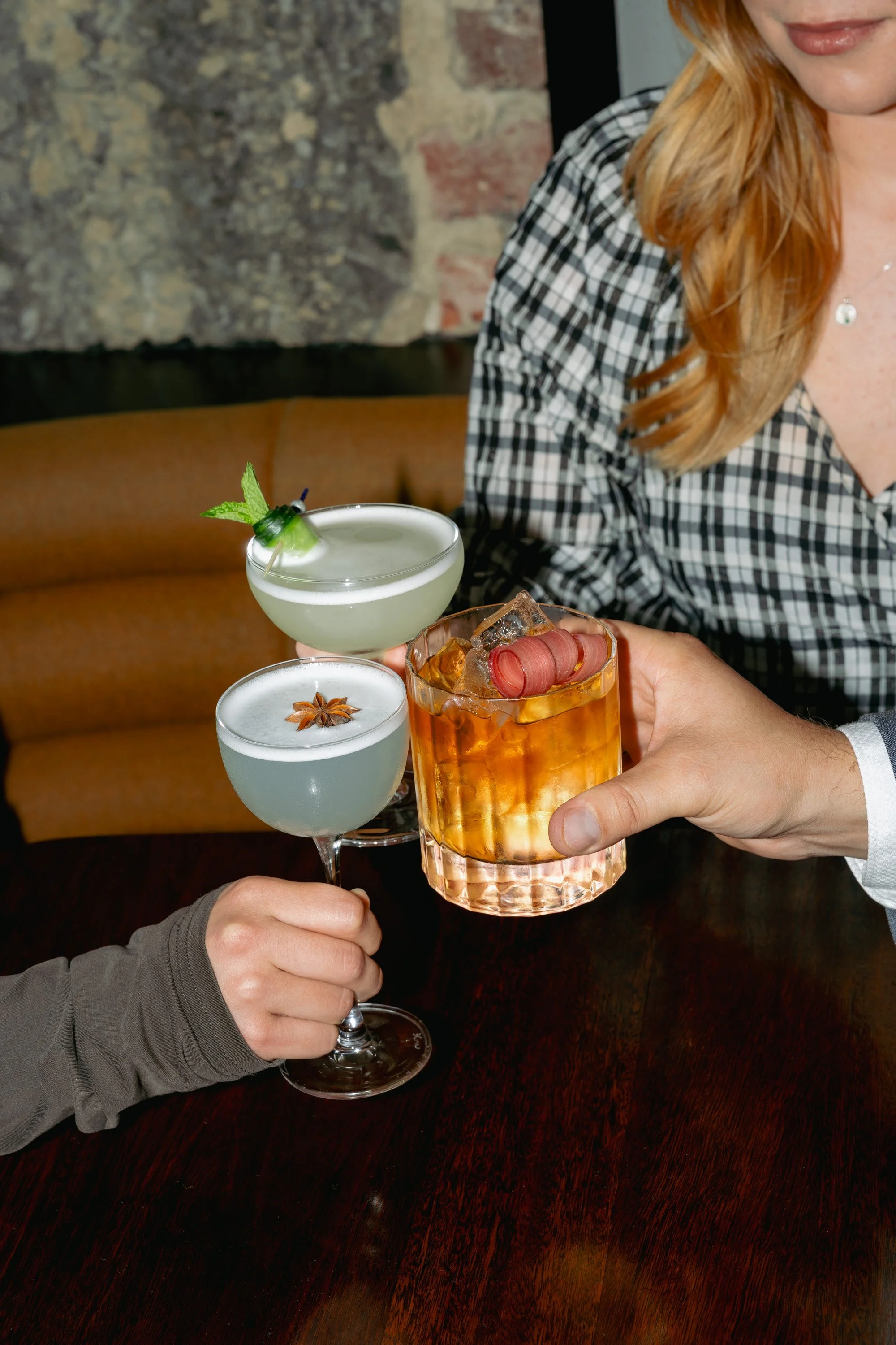 Three people clinking glasses containing colorful cocktails at a bar or restaurant.