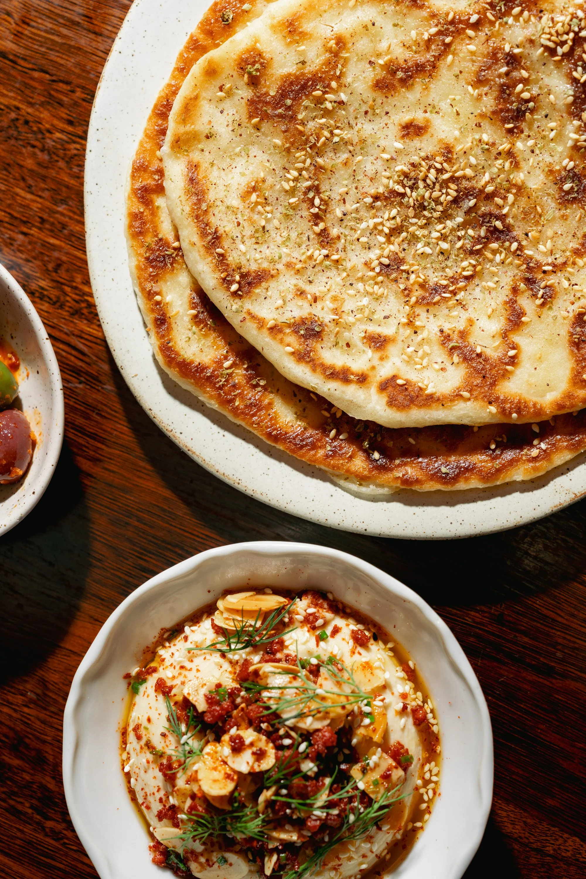 Two plates of Indian cuisine on a wooden table, featuring naan bread topped with sesame seeds and a bowl of curry with meat, vegetables, and herbs.