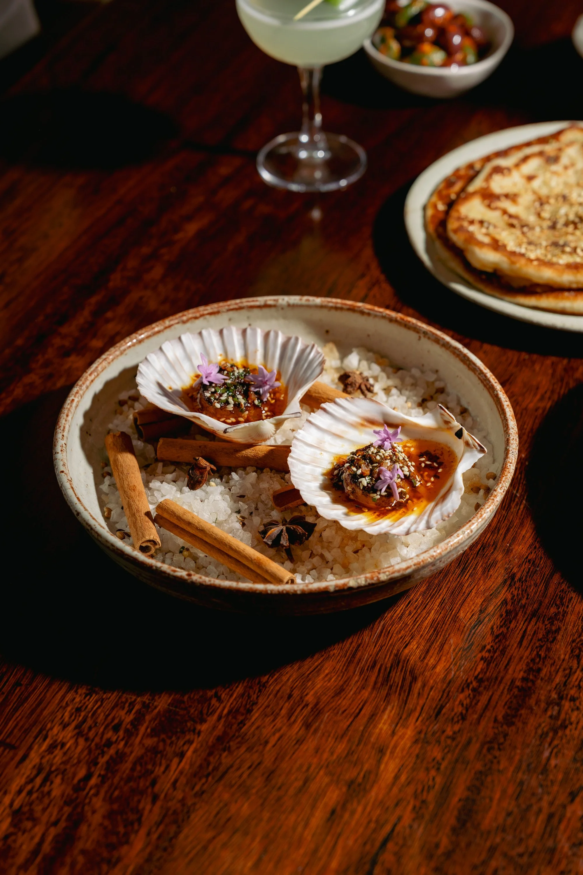 Seafood dish with scallops in shells garnished with herbs and flowers, served over sea salt with cinnamon sticks and star anise, on a rustic wooden table.