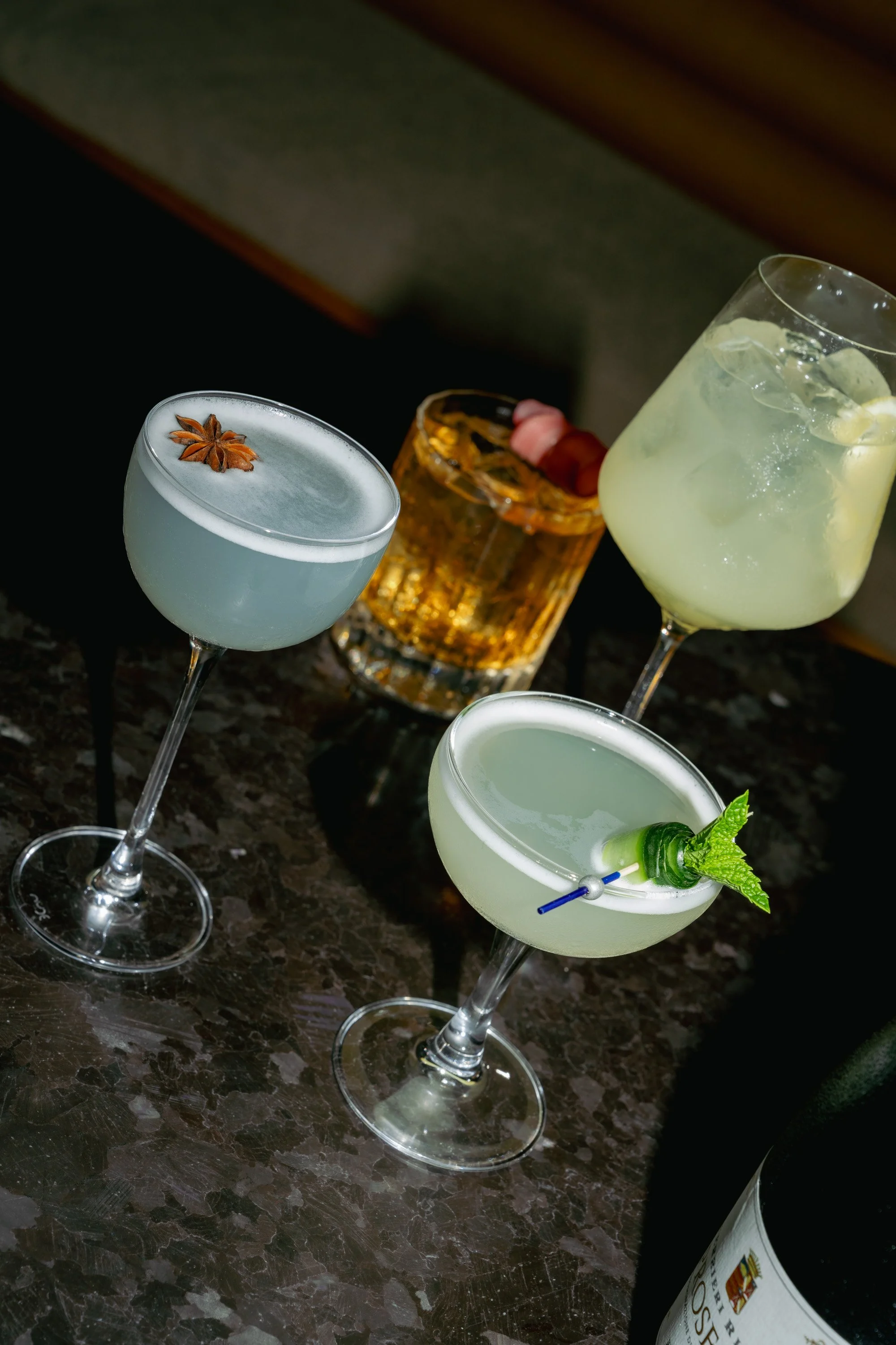 Four cocktails on a dark marble table, including a blue martini with a star anise garnish, a yellow cocktail with ice and lemon slices, a clear cocktail with mint and a straw, and a whiskey glass with ice and a lemon slice.