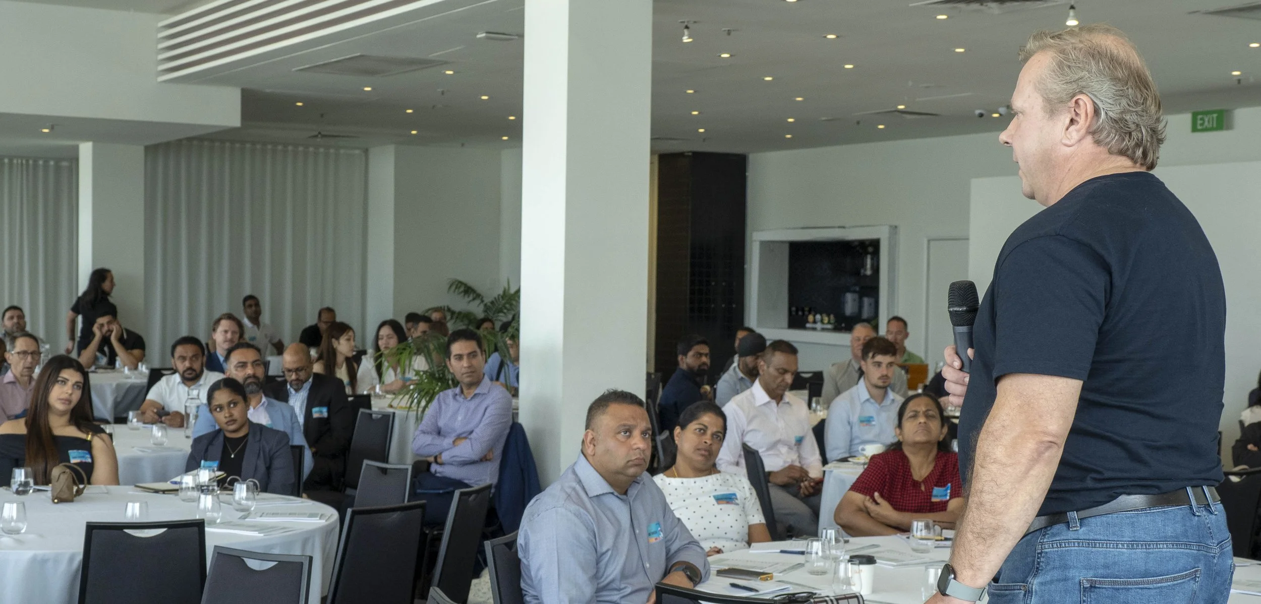 A speaker with gray hair, wearing a black T-shirt, holding a microphone, addressing an audience in a conference room with round tables and seated attendees listening attentively.