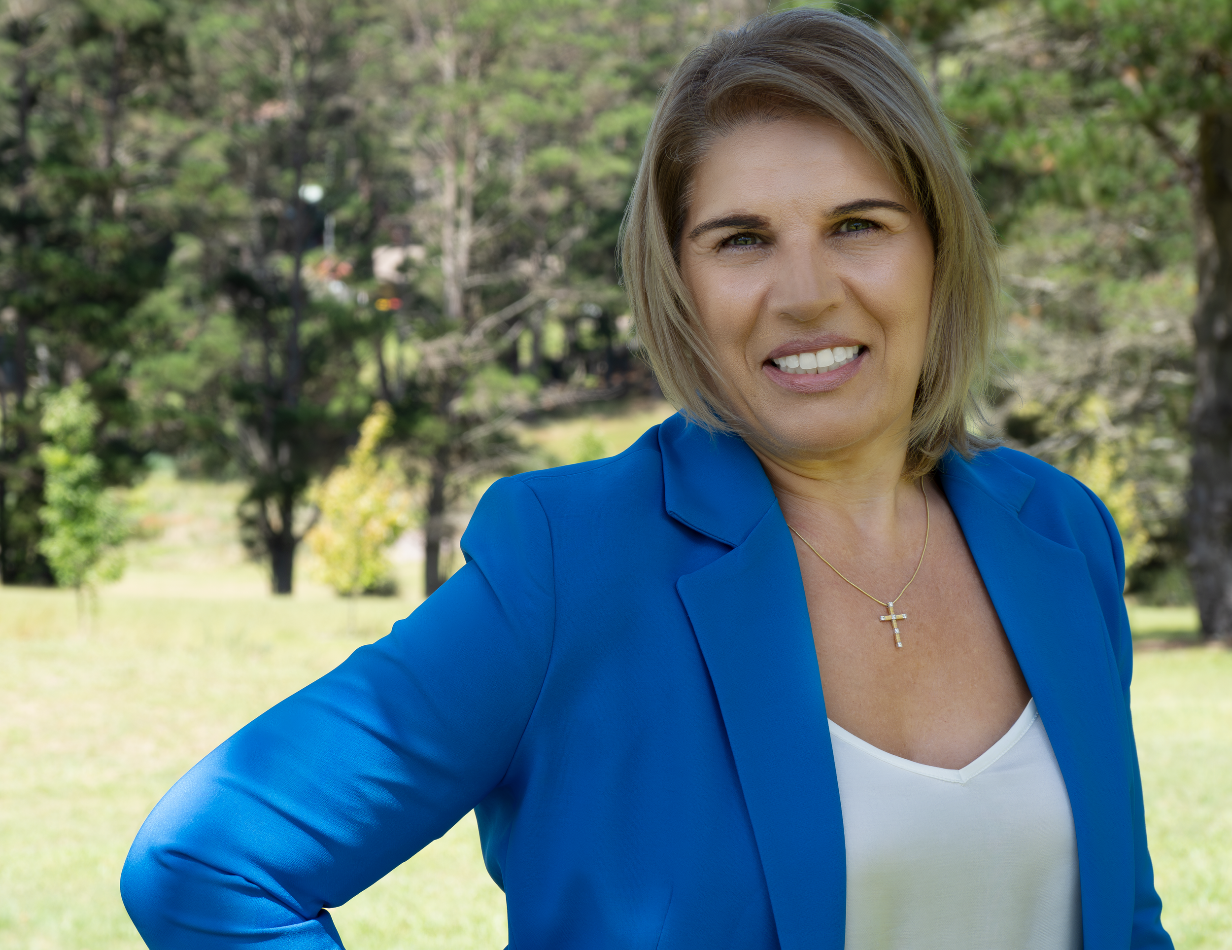 A middle-aged woman with blonde hair and blue eyes outdoors, wearing a blue blazer, white top, and a gold necklace with a cross pendant, standing in a park with trees in the background.