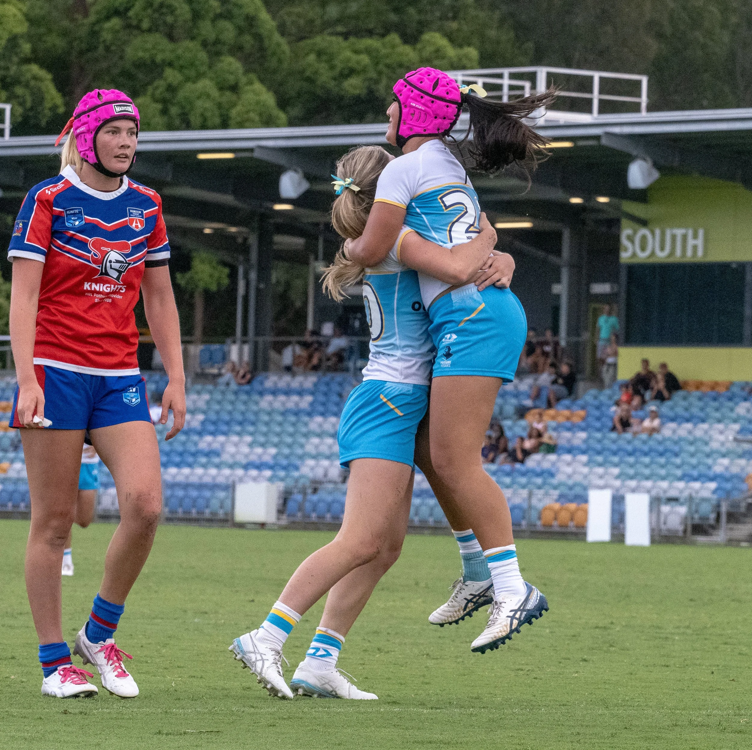 Two female athletes in cricket uniforms and pink helmets celebrate by hugging on a cricket field, with a third player watching nearby. In the background, there are empty stadium seats and a sign that reads 'SOUTH'.