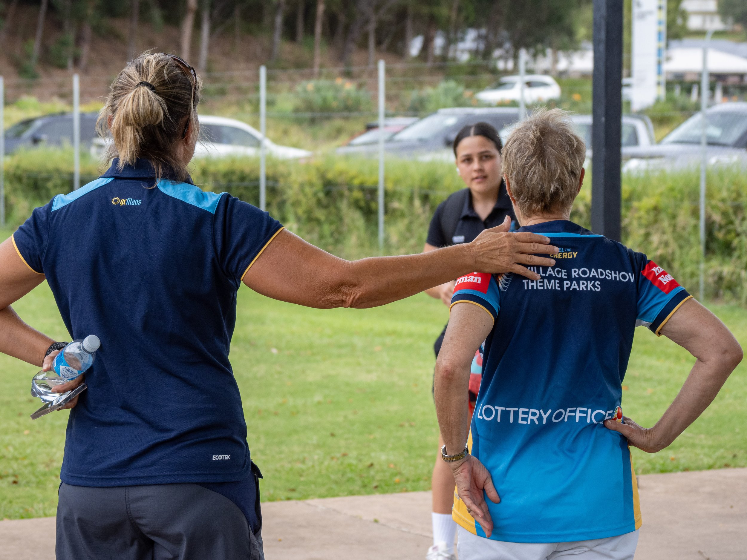 Women in sports uniforms talking outdoors, with one woman touching another woman's shoulder, in a grassy area near parked cars.