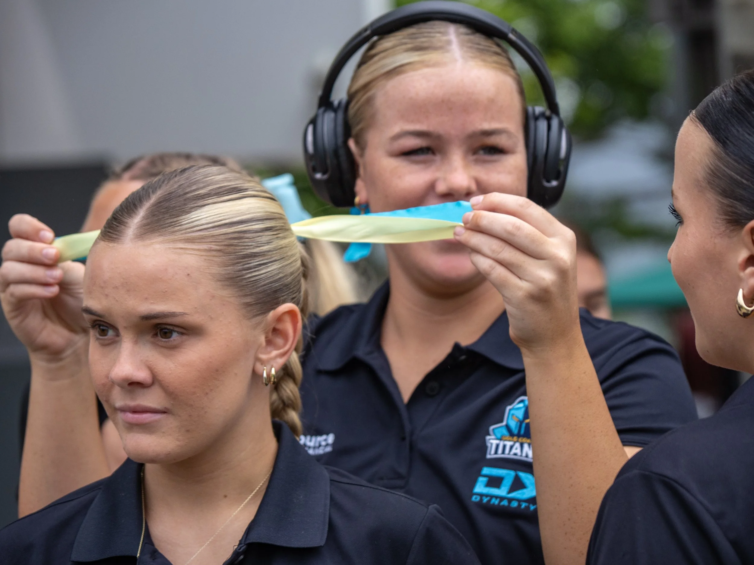A woman with headphones and a team uniform is holding a ribbon for a ceremonial ribbon cutting, while a girl with braided hair wearing a team uniform looks on.