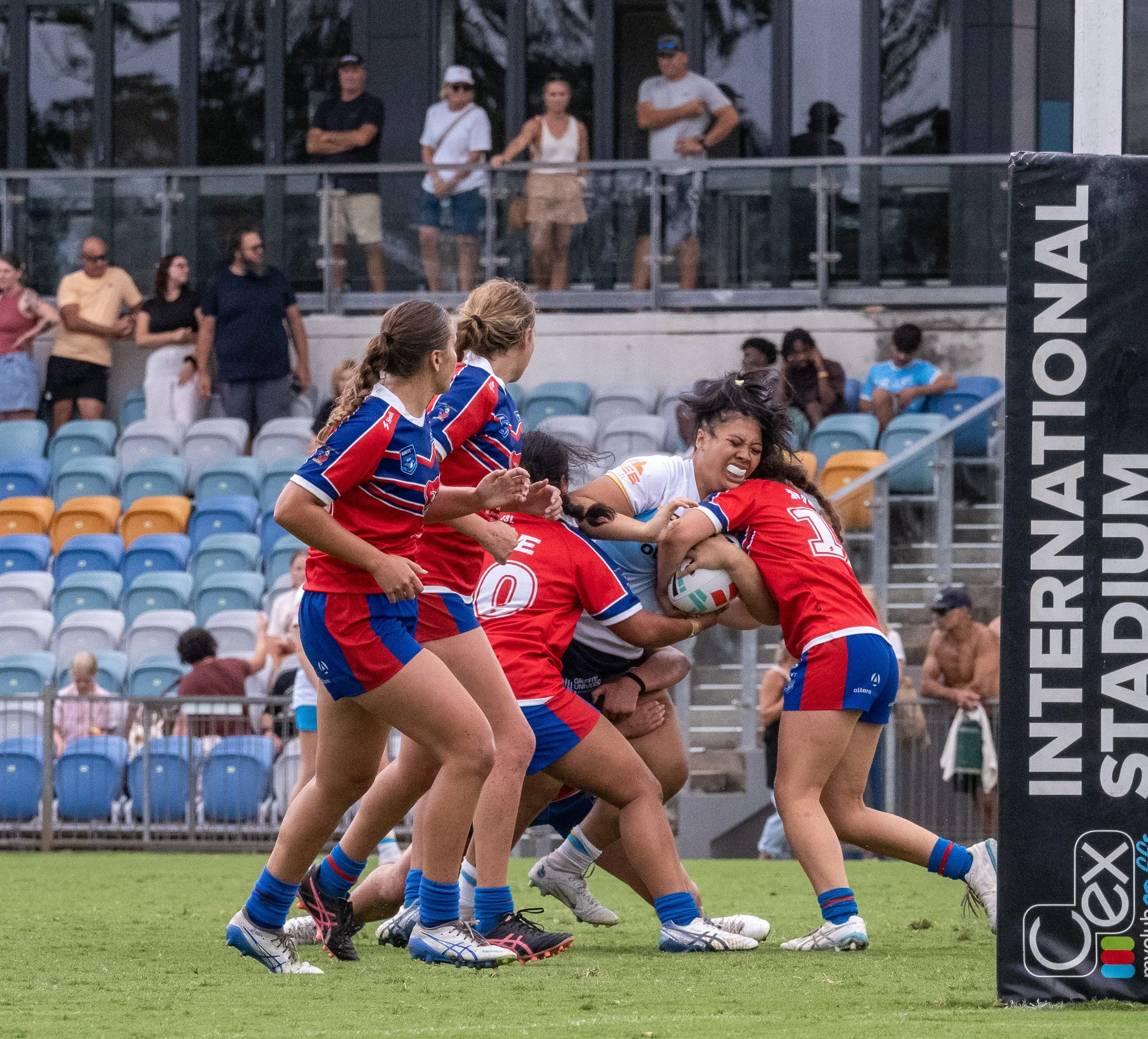 Women playing rugby, fighting for the ball near the goalpost, with spectators watching from the stands.