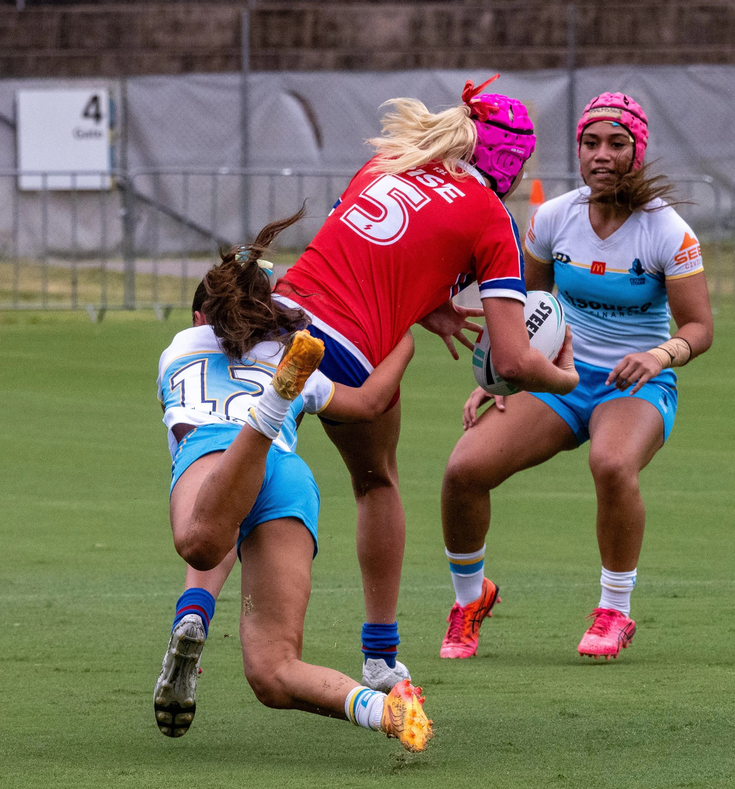 Three female rugby players competing for the ball during a game. Two players wear light blue and white uniforms, and one wears a red and blue uniform. One of the players in blue is attempting to tackle the player in red, while the other in blue stand