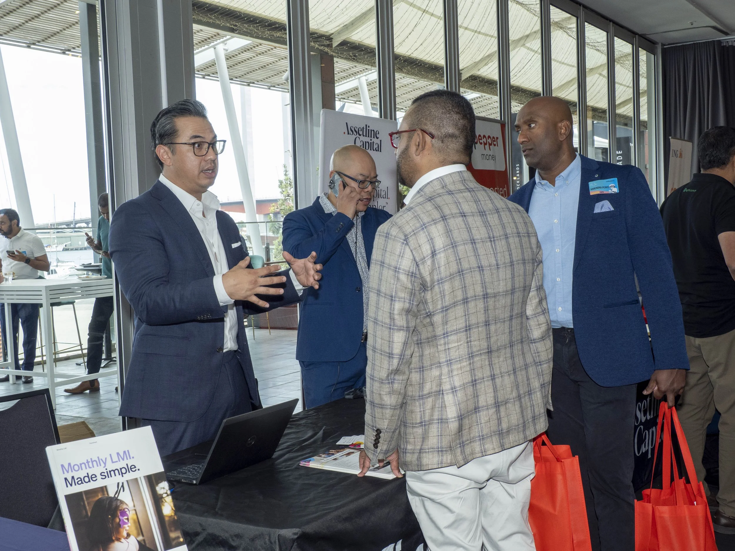 Business professionals engaging in conversation at a conference or trade show, with informational booths and banners in the background.