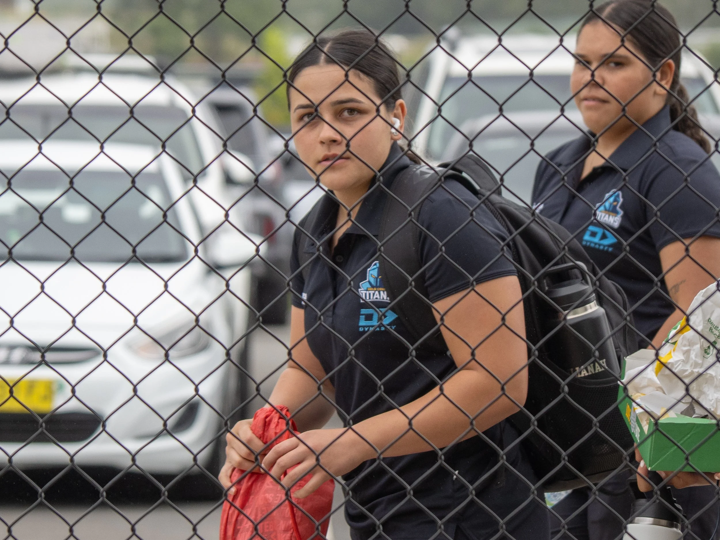 Two young women wearing black sports uniforms with a volleyball team logo, standing behind a chain-link fence in a parking lot.