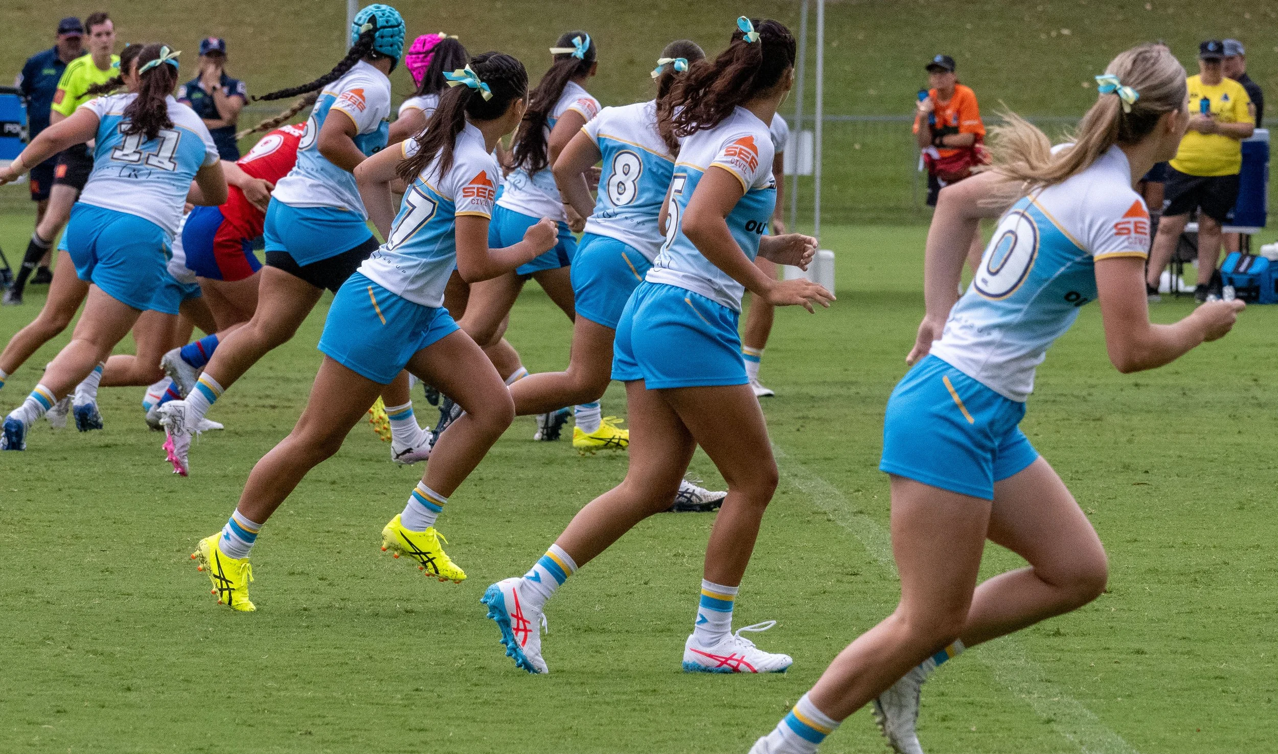 Group of female athletes, likely rugby players, running on a grassy field during a match or practice. They are dressed in matching blue and white uniforms with numbers and logos, wearing cleats and athletic socks, with some wearing headbands or bows.