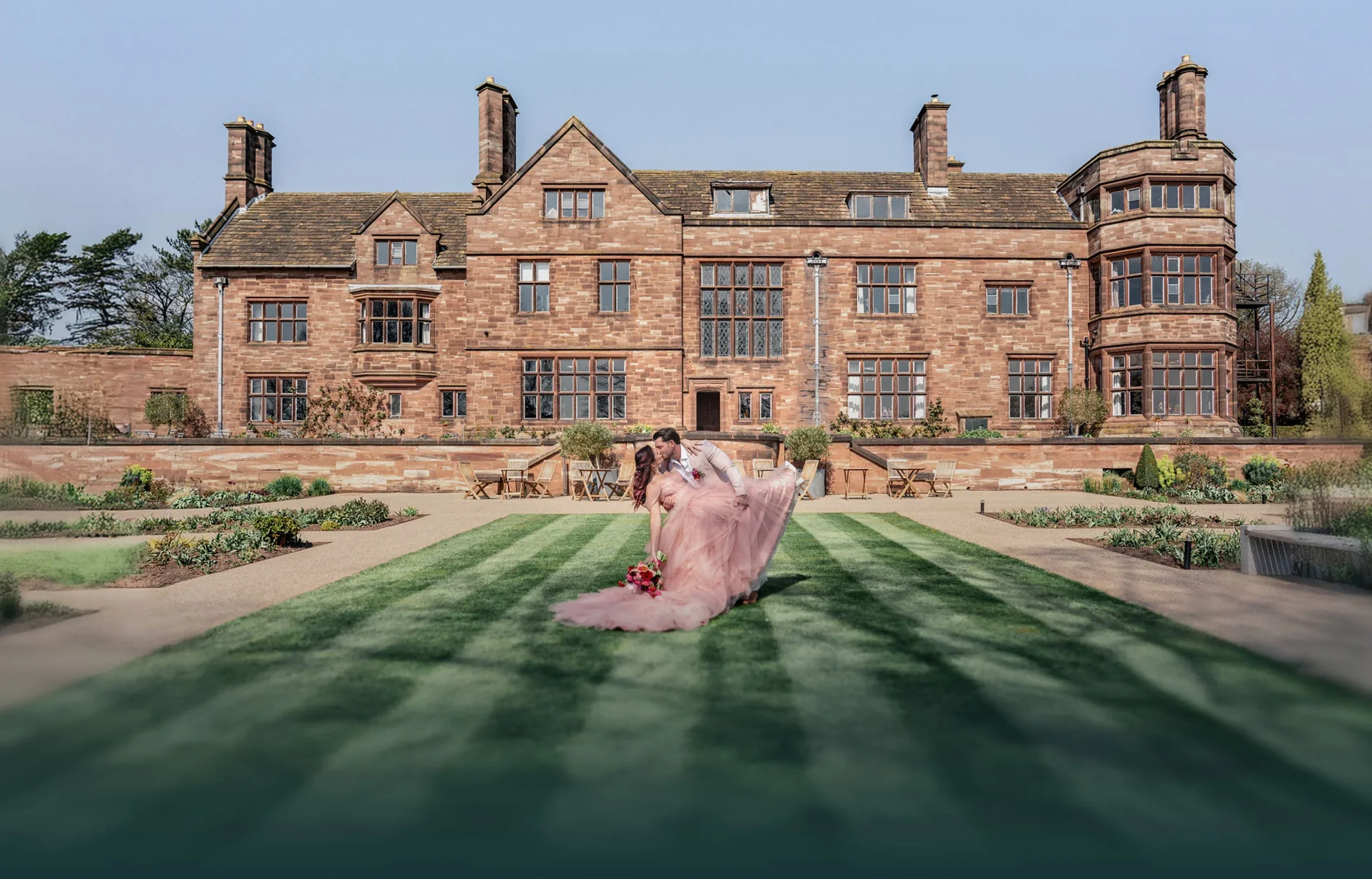 A couple dancing on a well-manicured lawn in front of a large, historic stone mansion, Salt House, Standon Hall wedding venue Eccleshall during daytime.