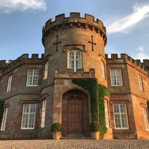 A stone castle with a central tower, arched wooden door, and multiple windows, some with decorative cross accents, surrounded by greenery and potted plants under a partly cloudy sky. Weddings at the citadel weston north shropshire