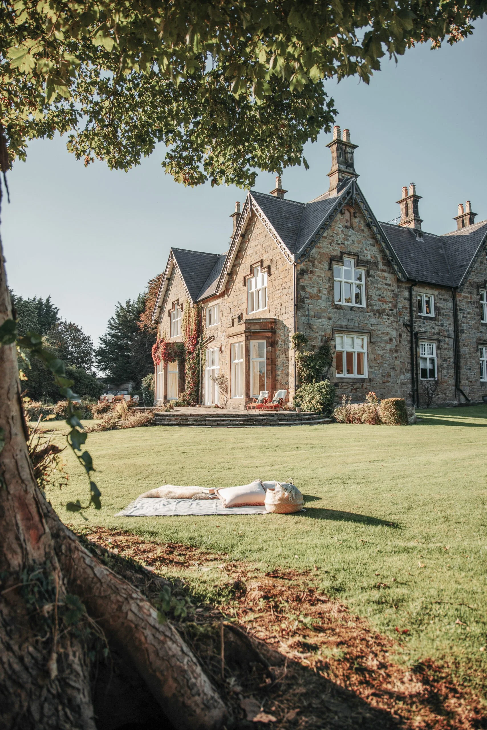 A large stone house with multiple chimneys and steep roof, surrounded by trees and a well-maintained lawn. Weddings at Ein Cynefin nr Mold luxury private dining and holiday let