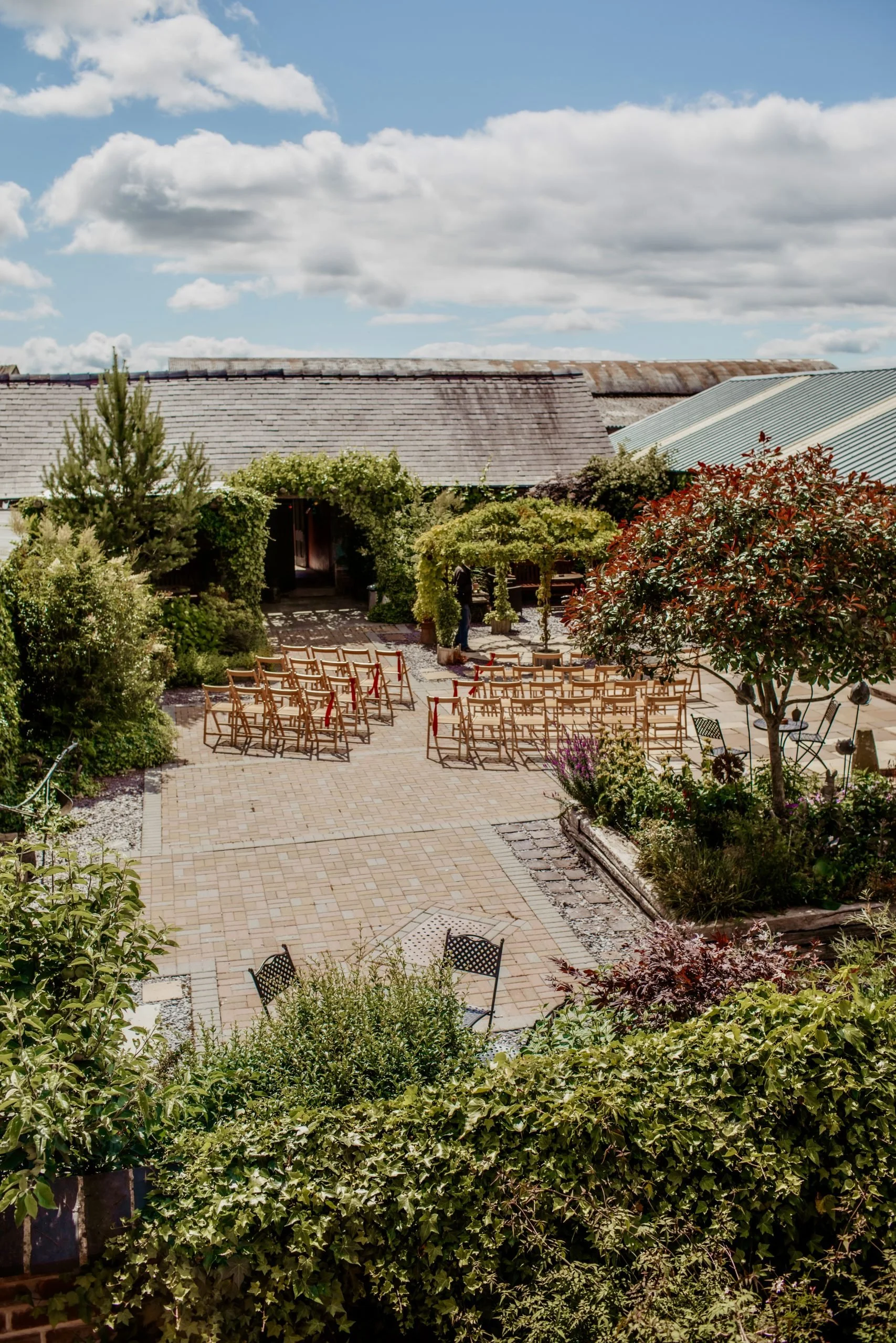 A cozy outdoor garden with seating, lush greenery, flowering plants, and a partly cloudy sky. Wedding at Bromwich park oswestry