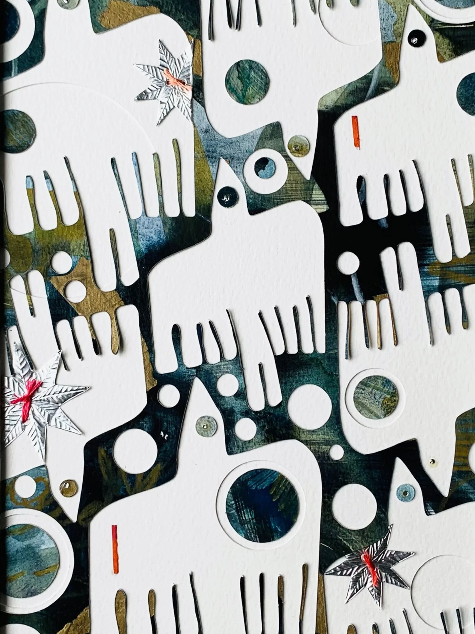 Close-up of a decorative art piece with cut-out shapes, some with reflective surfaces, and small embellishments including paper stars with red ties, on a textured white background.