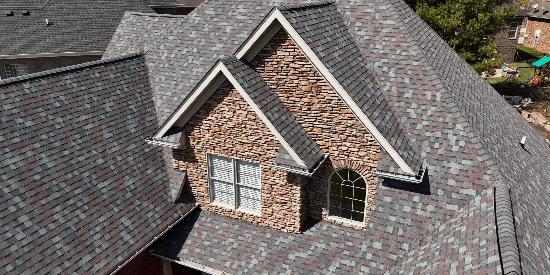 Aerial view of a house roof with new roof, featuring gray and red asphalt shingles and a brick chimney with stone accents, along with two windows and surrounding trees and neighboring homes.