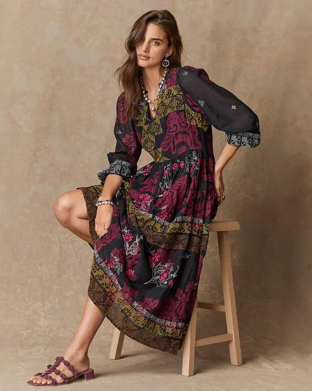 A woman with wavy brown hair wearing a colorful patterned maxi dress, accessorized with earrings, a necklace, and bracelet, sitting on a wooden stool against a beige background.