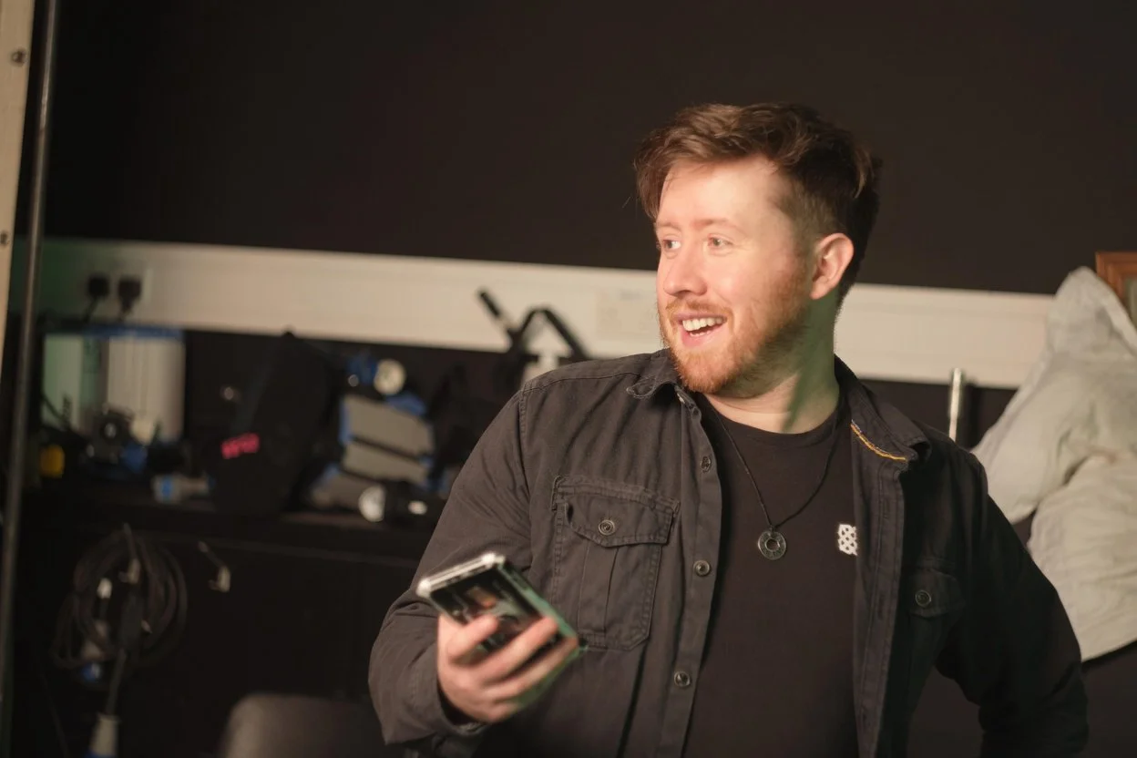 A young man with light brown hair and a beard, smiling and holding a remote control, sitting on a bed in a bedroom with a black wall and various electronic equipment in the background.