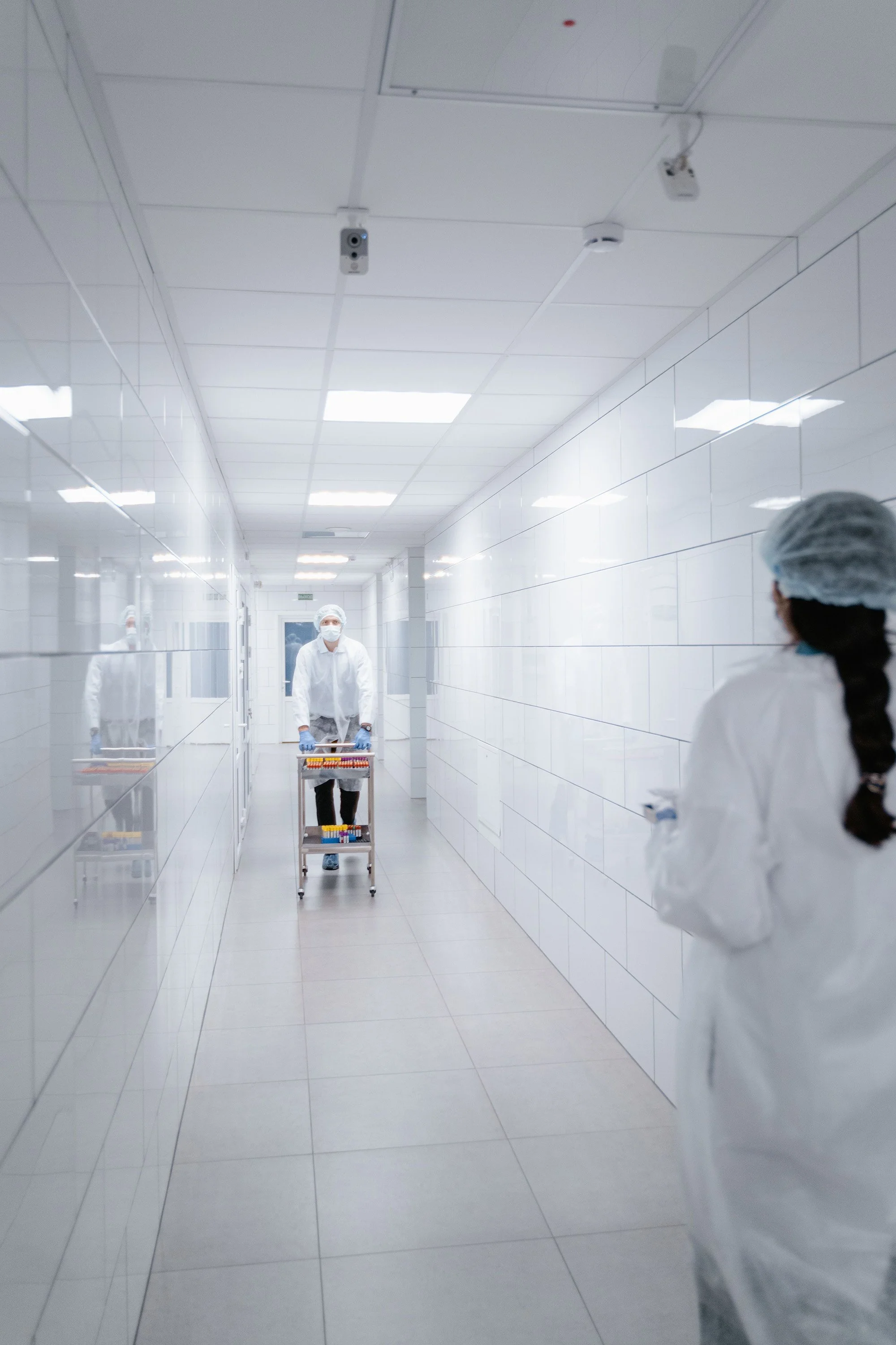 A person dressed in medical attire, including a mask, gloves, and hair cover, pushes a cart with test tubes down a brightly lit, white-tiled hallway in a medical or laboratory setting. Two other figures in similar attire are visible in the hallway.