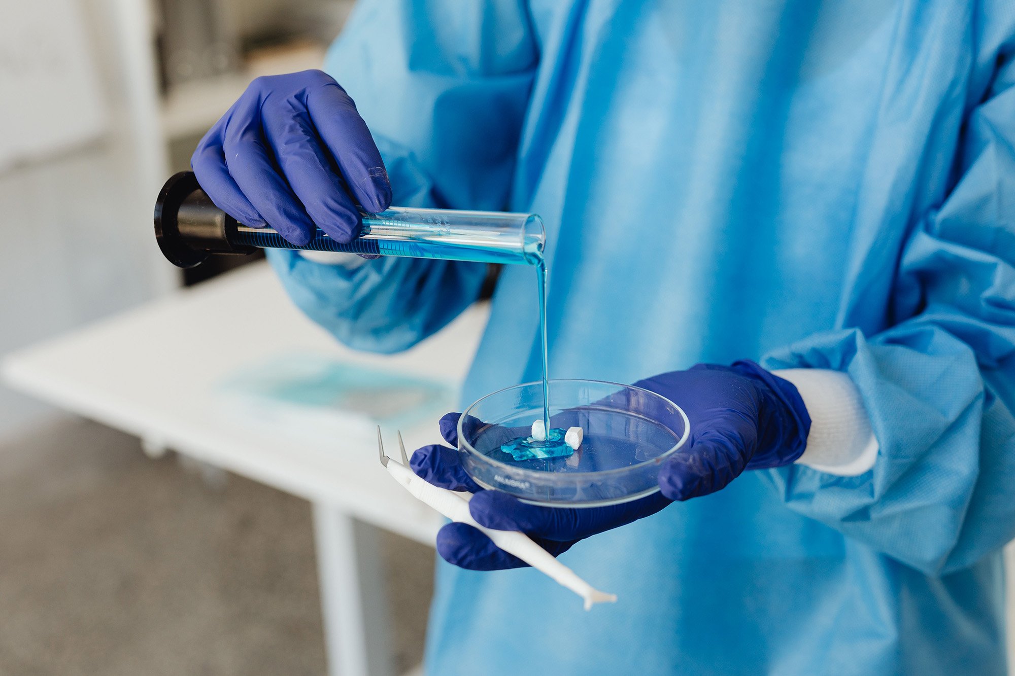 A scientist wearing blue gloves and a blue protective gown pouring a blue liquid into a petri dish.