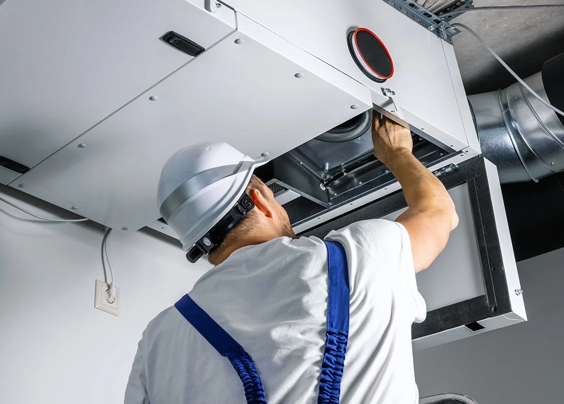 Worker wearing a white safety helmet and white shirt with blue suspenders installing or repairing a HVAC unit on the ceiling.
