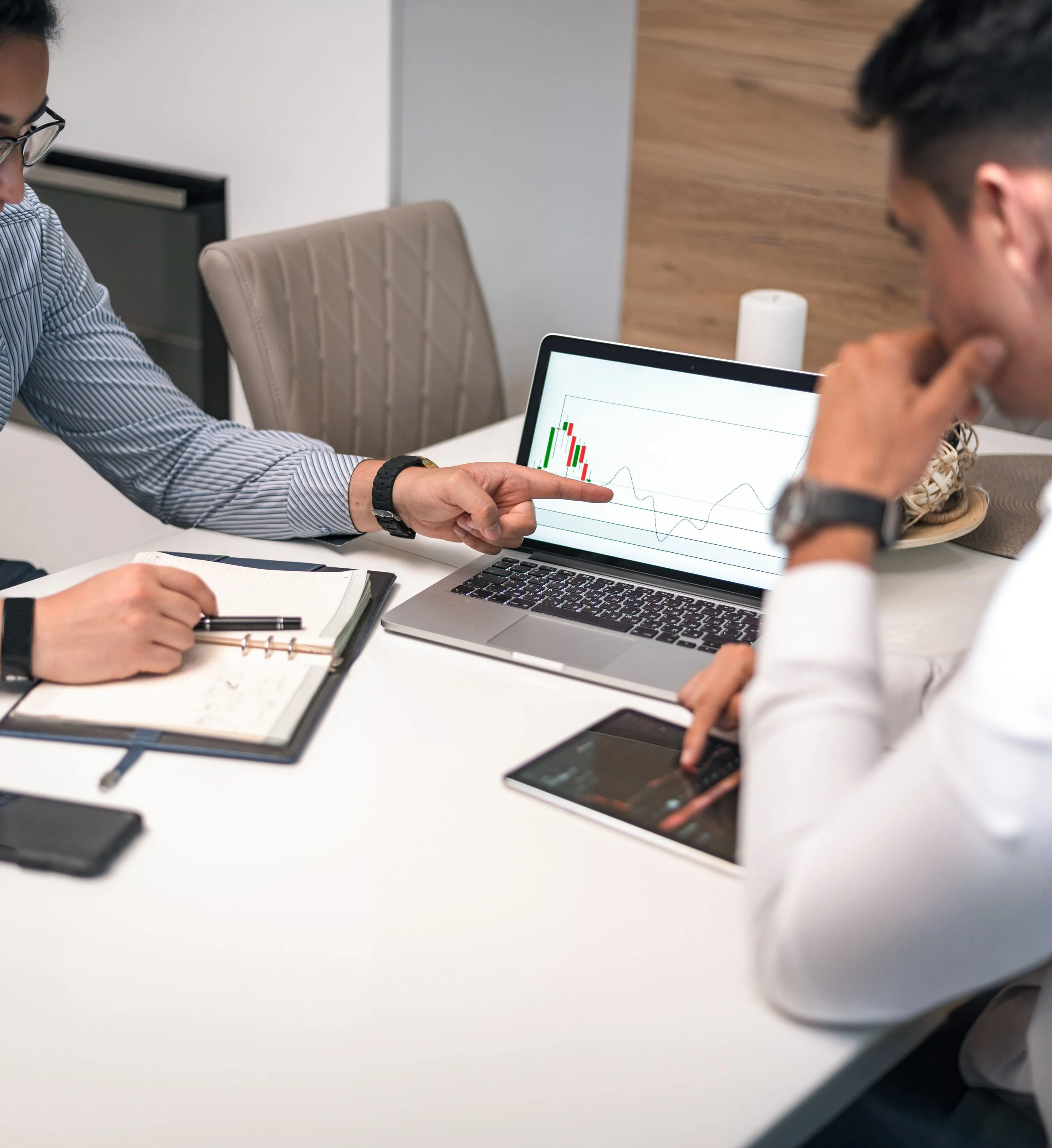 Two individuals are engaged in a discussion, with one pointing at a laptop screen displaying a graph with red and green candlesticks and a line chart. The table also has notebooks, a pen, a smartphone, and a smart speaker. The setting appears to be a modern office or meeting room with beige and wooden decor.