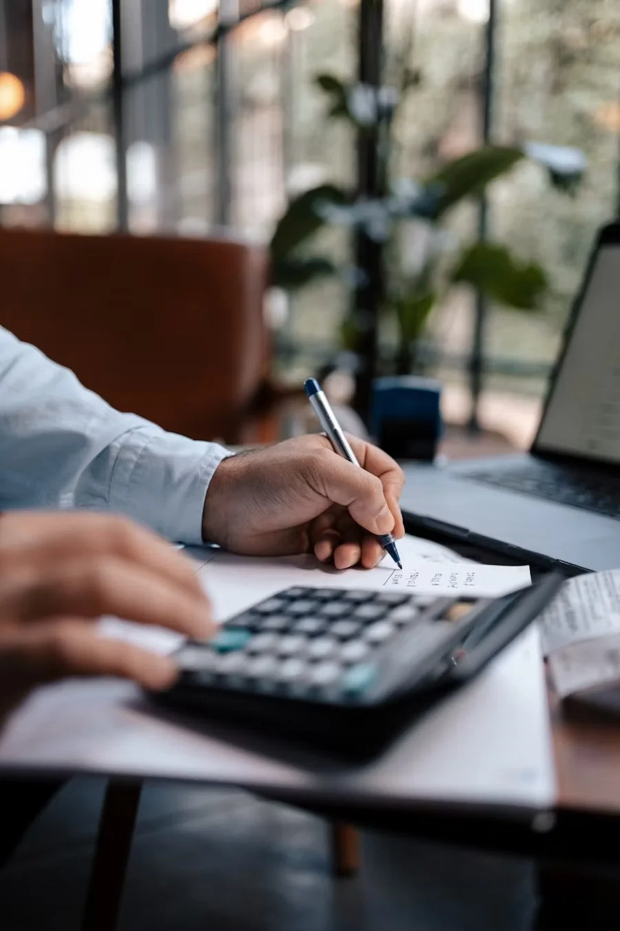 A person is sitting at a desk, using a calculator and writing on a piece of paper with a pen. There is a laptop and some documents on the desk, with a blurred background of a window and tree outside.