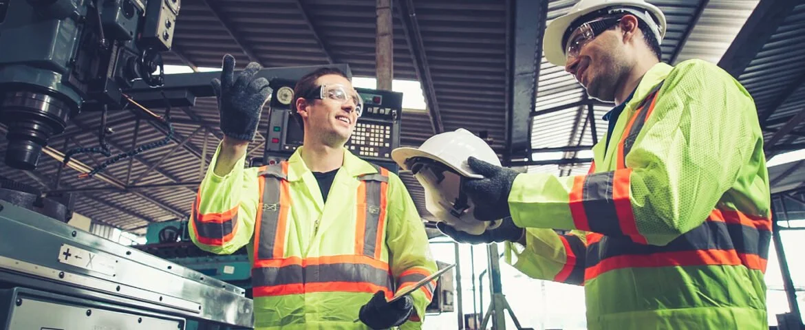 Two construction workers wearing high-visibility yellow and orange safety vests, helmets, and gloves inside an industrial building, engaged in a discussion, with one holding a clipboard and the other taking notes on a tablet.