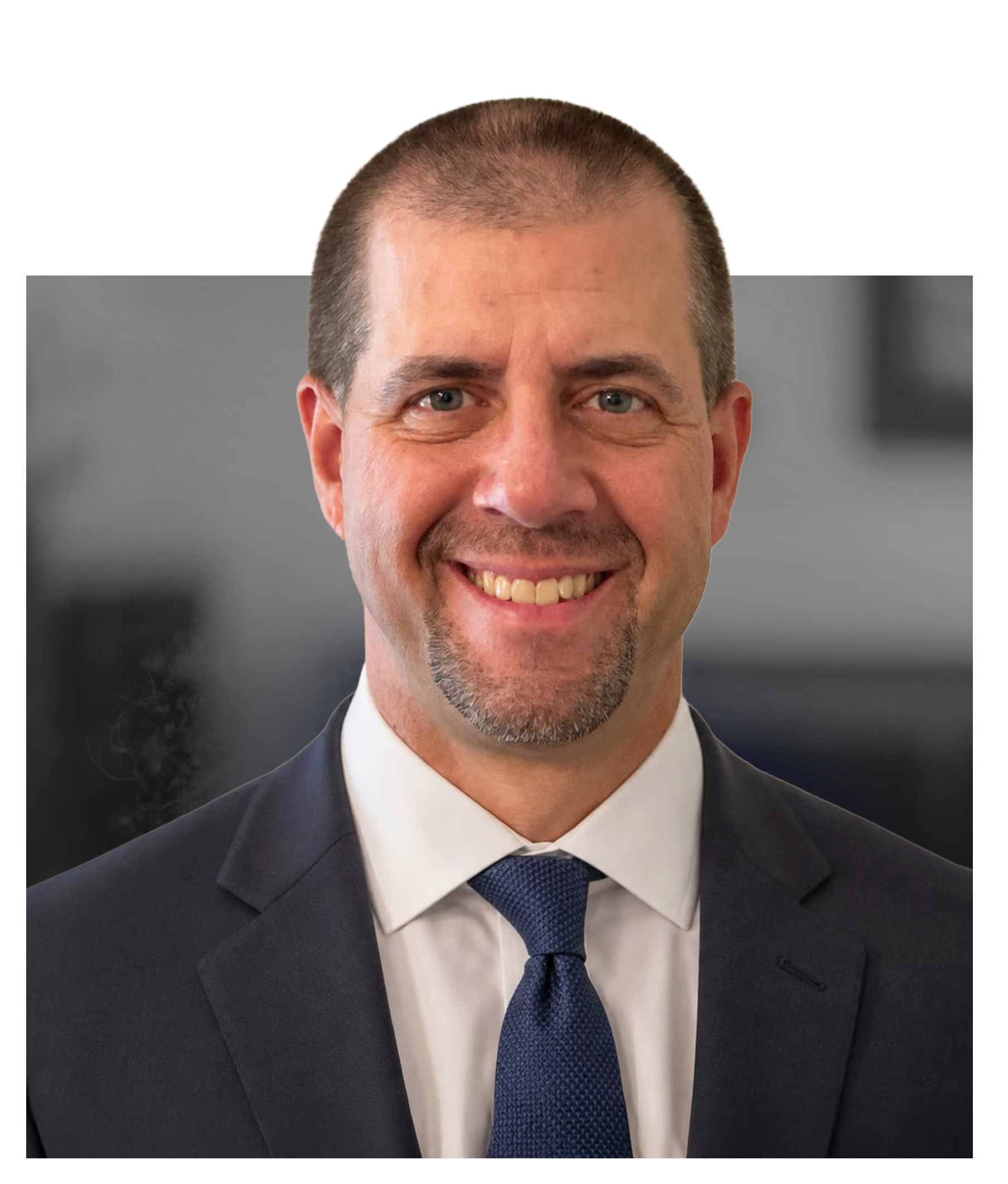 Close-up of a smiling man wearing a dark suit, white shirt, and blue tie, with short hair and a trimmed beard.