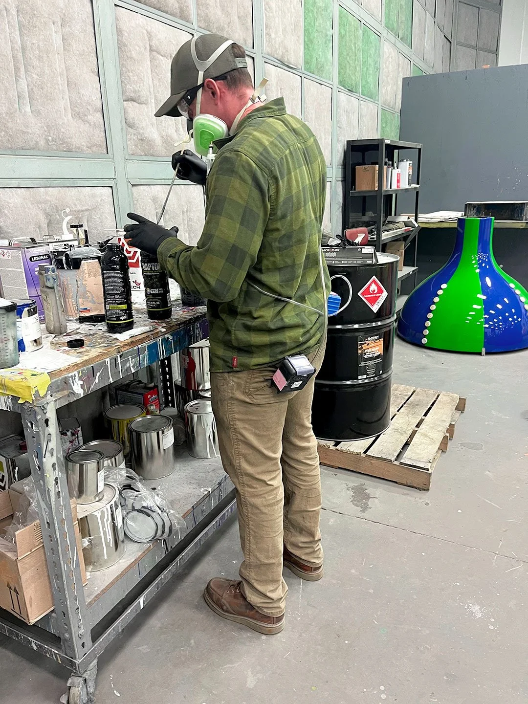 A man in a green plaid shirt, tan pants, and brown shoes inspecting paint cans in a workshop, wearing a face mask, gloves, and a gray cap.
