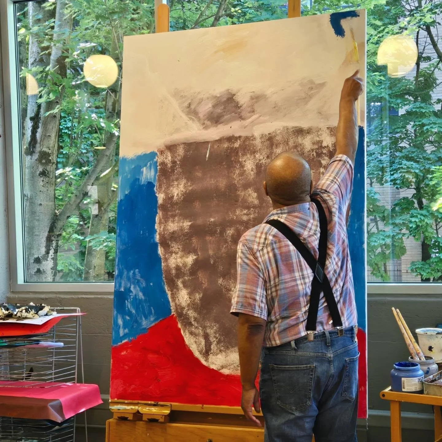 Happy friday! Big things happening in the studio this week ✨️ 

ID: Artist Mark Bishop standing facing a large canvas on an easel. On the canvas there is a work in progress painting with blue, red, brown, and white colors visible. Artist is holding a