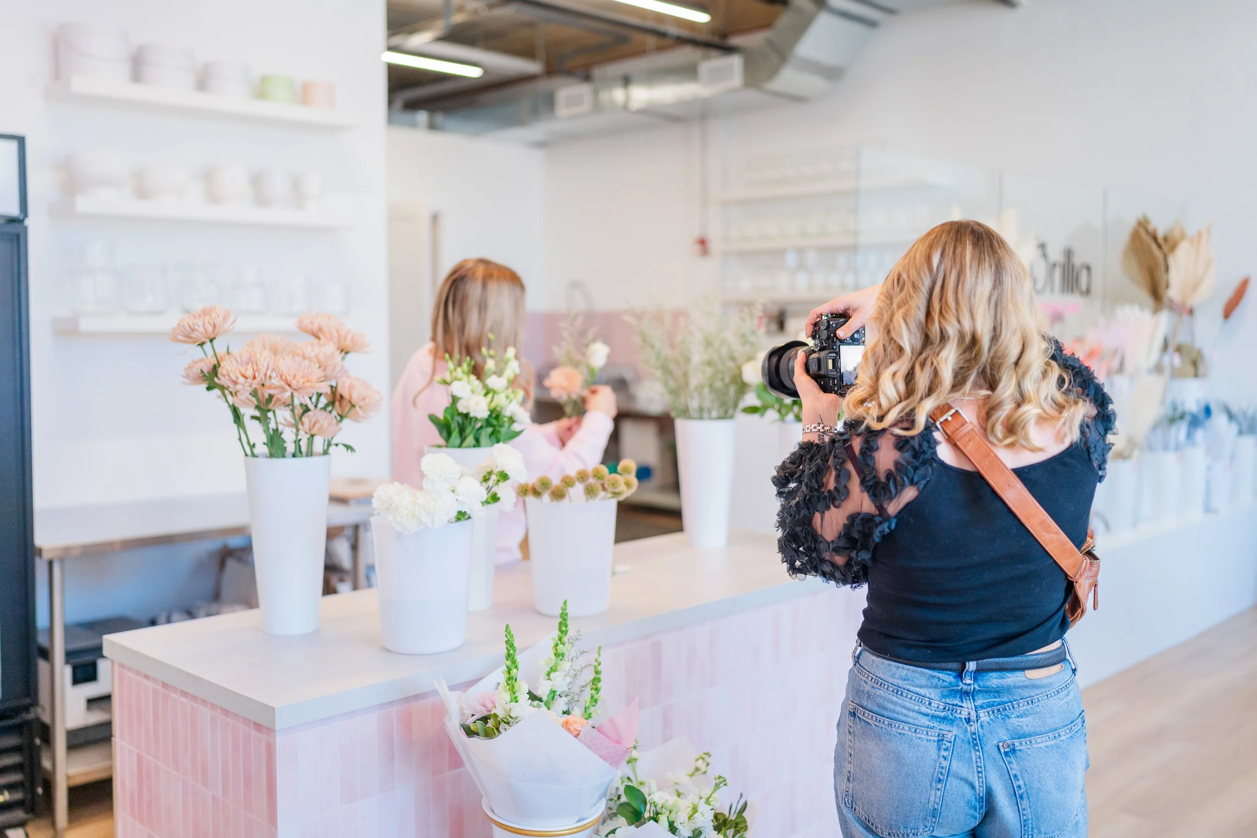 Personal brand photographer is photographing a floral arrangement in a flower shop. The shop has various vases of flowers on a counter and shelves with decor items in the background. Another person is arranging flowers behind the counter.